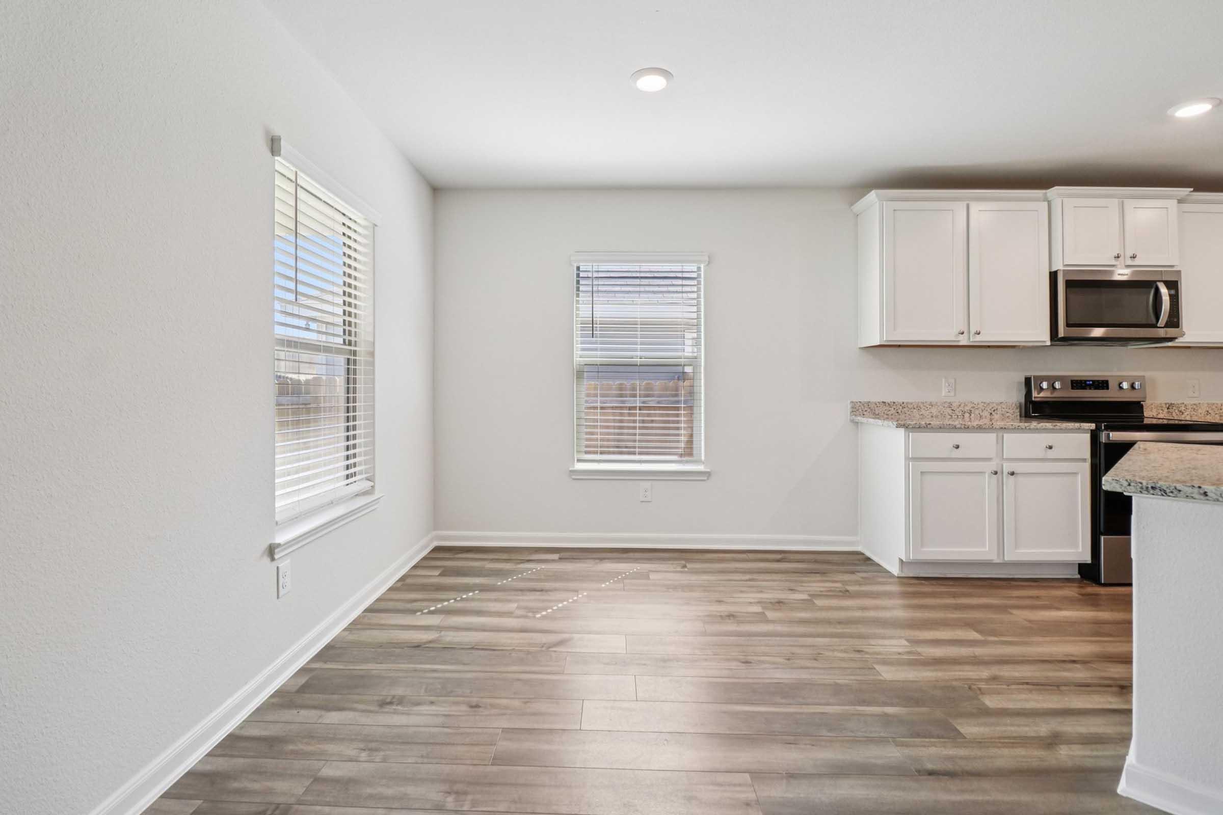 An empty, modern kitchen and living area featuring light-colored walls, a window with blinds, and wood-like flooring. The kitchen has white cabinets, a stainless steel microwave, and a countertop. Natural light brightens the space.