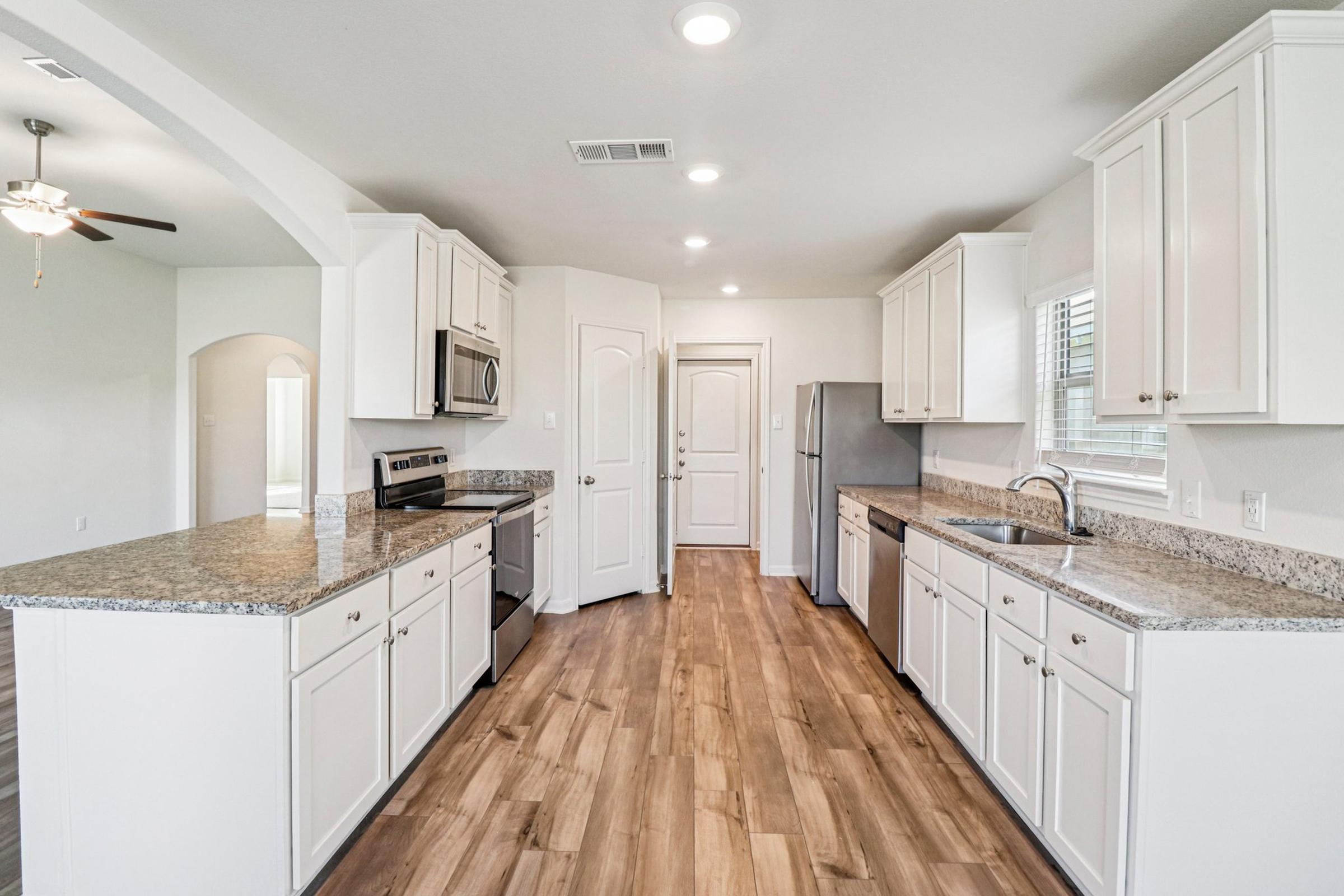 A modern kitchen featuring white cabinetry, stainless steel appliances, and a large granite countertop island. The space has wood-style flooring, large windows for natural light, and an open layout connecting to another room.