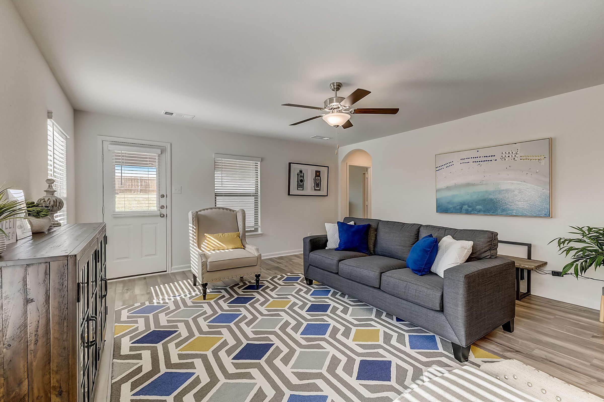 A cozy living room featuring a gray couch with blue cushions, a beige accent chair, and a stylish rug with geometric patterns. Bright light streams through the windows, illuminating the space. A wooden console table and decorative plant add to the inviting atmosphere, with a ceiling fan for added comfort.
