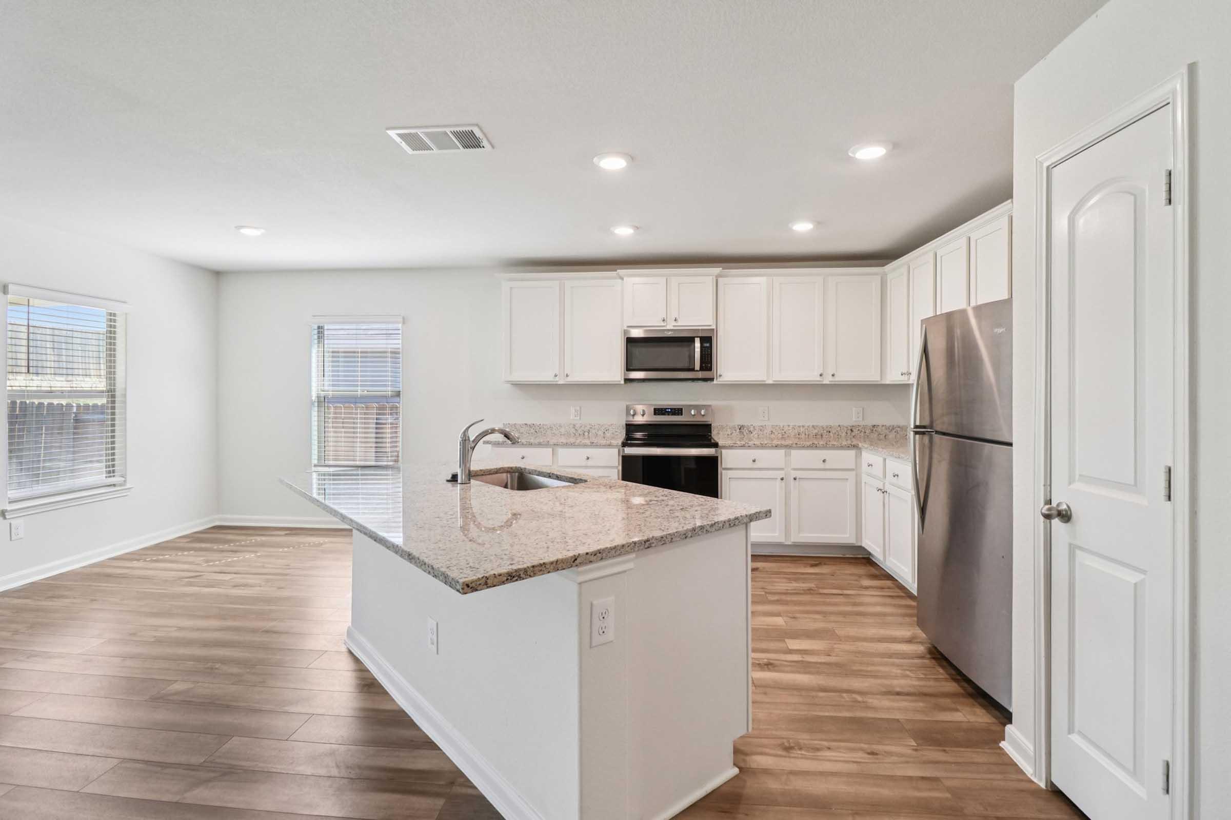 Modern kitchen featuring white cabinetry, stainless steel appliances, a granite countertop island, and hardwood flooring. Natural light floods the space through windows, highlighting the open layout.