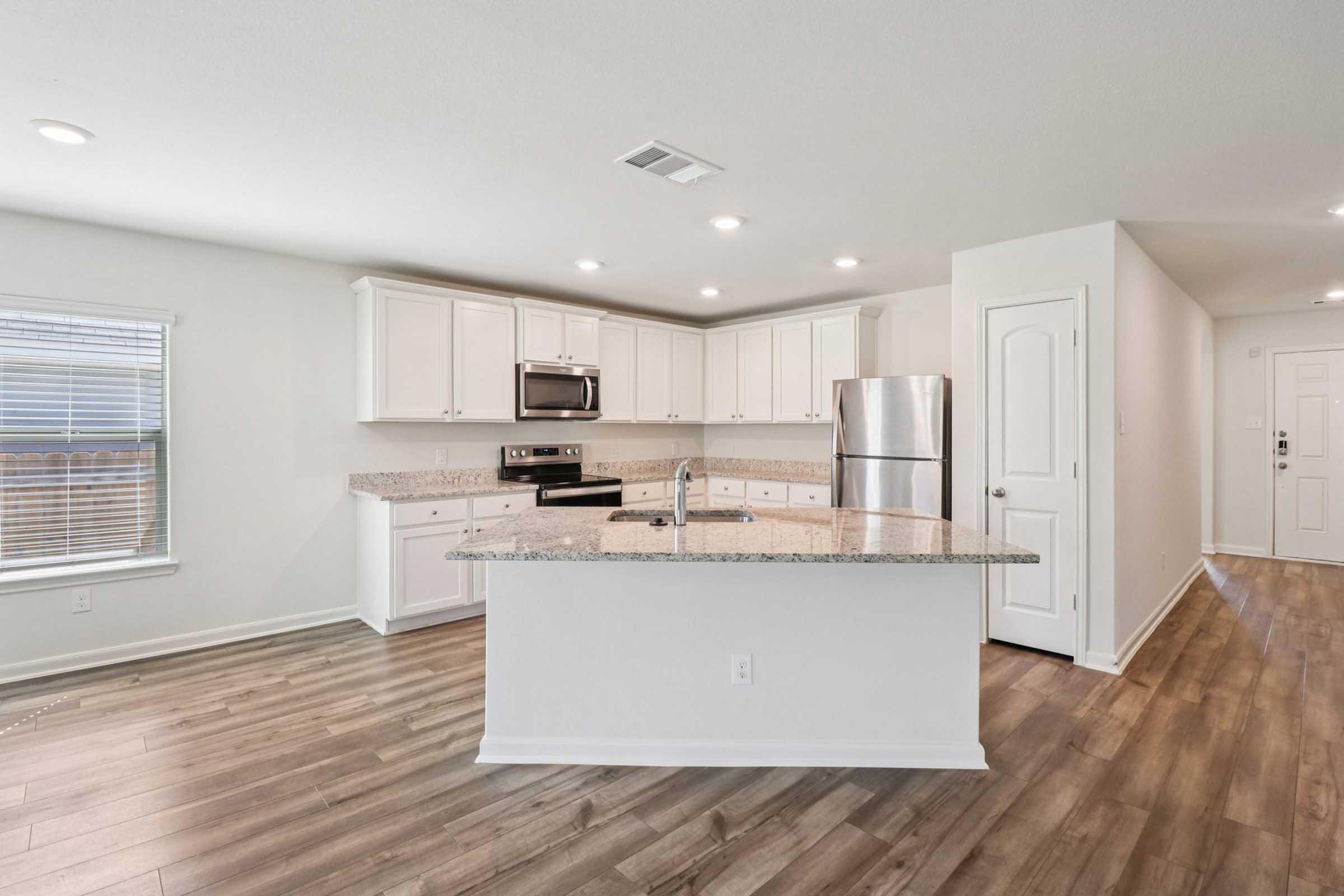Bright, modern kitchen featuring white cabinets, stainless steel appliances, and a spacious island with a granite countertop. The room is well-lit with recessed lighting and a large window. The flooring is wood-like, adding warmth to the space, and there's an entrance visible on the right side.