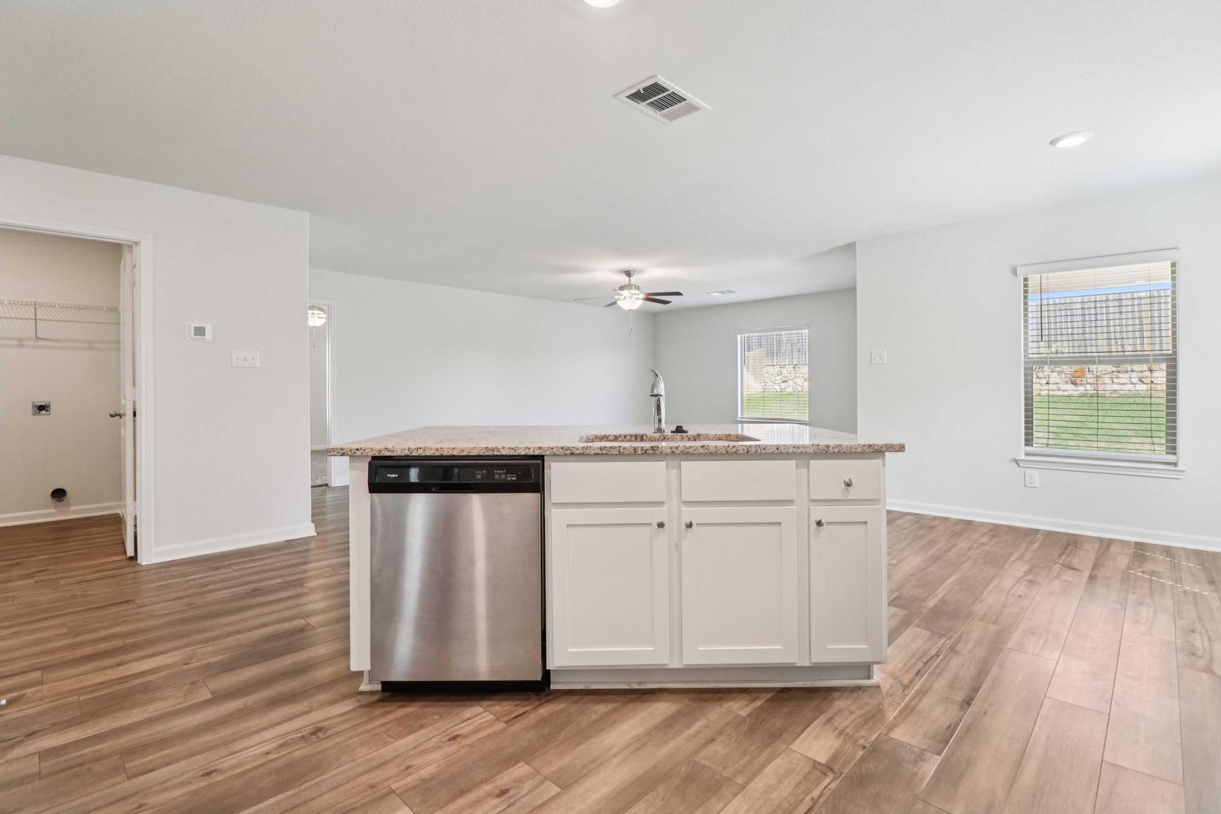 A modern kitchen featuring a central island with a granite countertop, a stainless steel dishwasher, and white cabinetry. The open floor plan connects to a well-lit living area with natural light, hardwood flooring, and neutral-colored walls. A door leads to a closet space.
