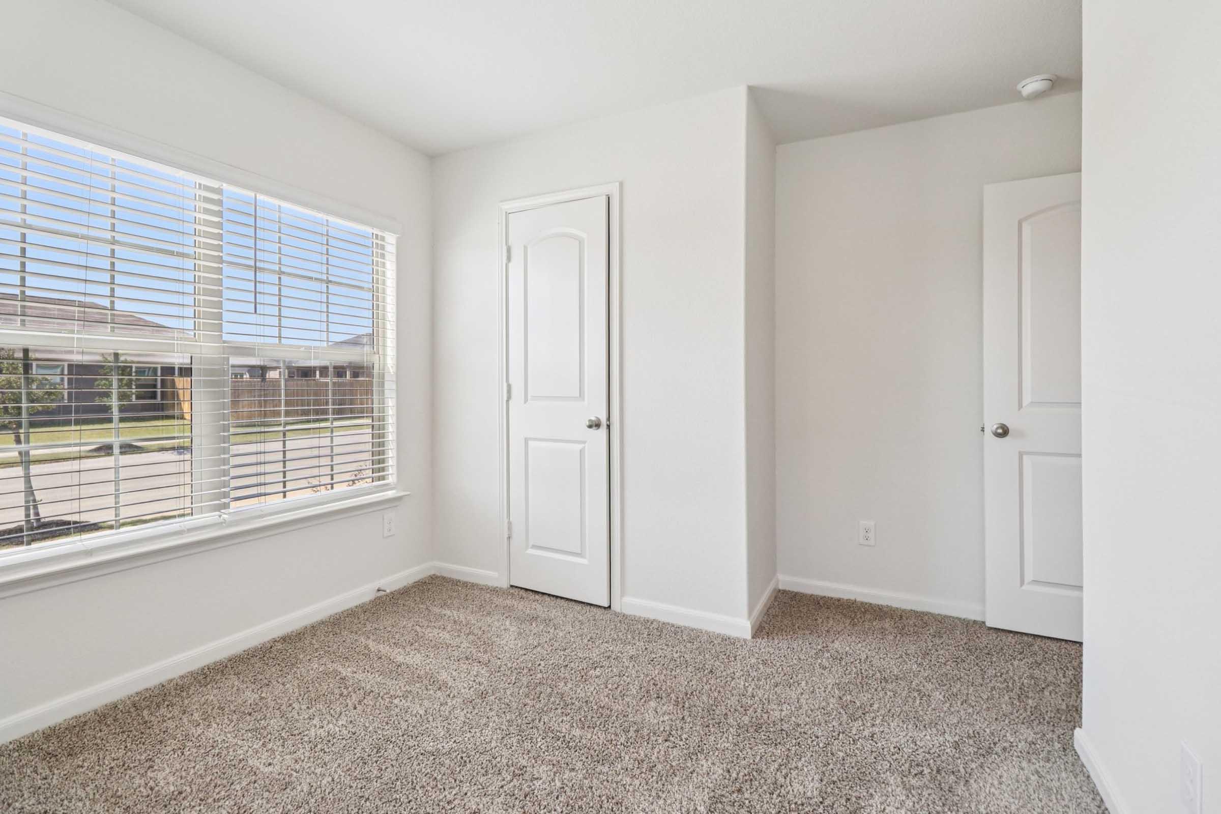 A bright, empty room featuring light-colored walls and plush carpet. A large window with blinds allows natural light to fill the space, showing a view of the outdoors. There are two doors; one leading to another room and the other likely to a closet. The overall ambiance is clean and spacious.
