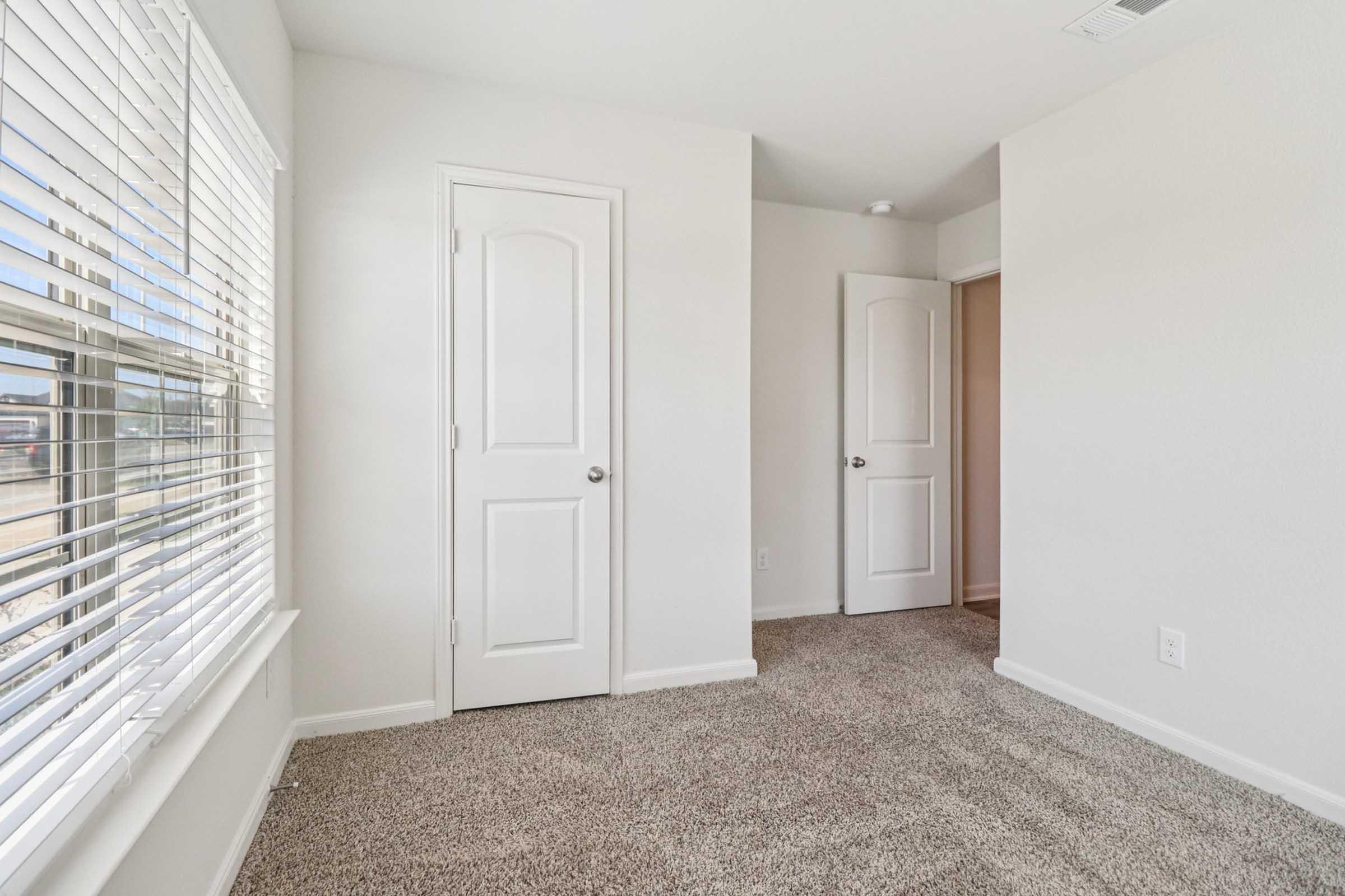 A bright, empty room with light-colored walls and plush carpet. Two white doors lead to adjacent spaces, and a window with blinds allows natural light to fill the area. The overall atmosphere is clean and spacious.