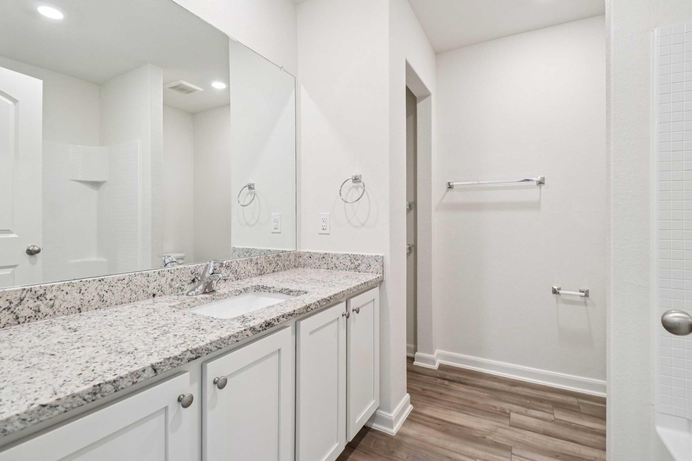 A modern bathroom featuring a granite countertop and sink with white cabinetry. The space includes a large mirror above the sink, a towel rack on the wall, and a walk-in shower visible in the background. Light-colored walls and wooden flooring contribute to a clean and spacious look.