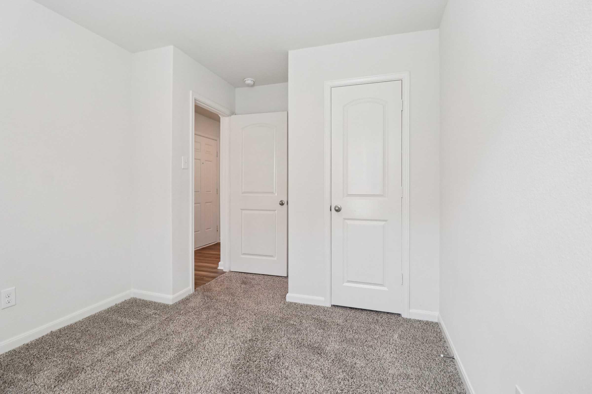 A small room with light-colored walls and plush gray carpet. There are two doors: one leading to an adjacent room and another closet door. The doorways are framed with simple white trim, creating a clean and minimalistic look. Natural light may be coming from an unseen window.