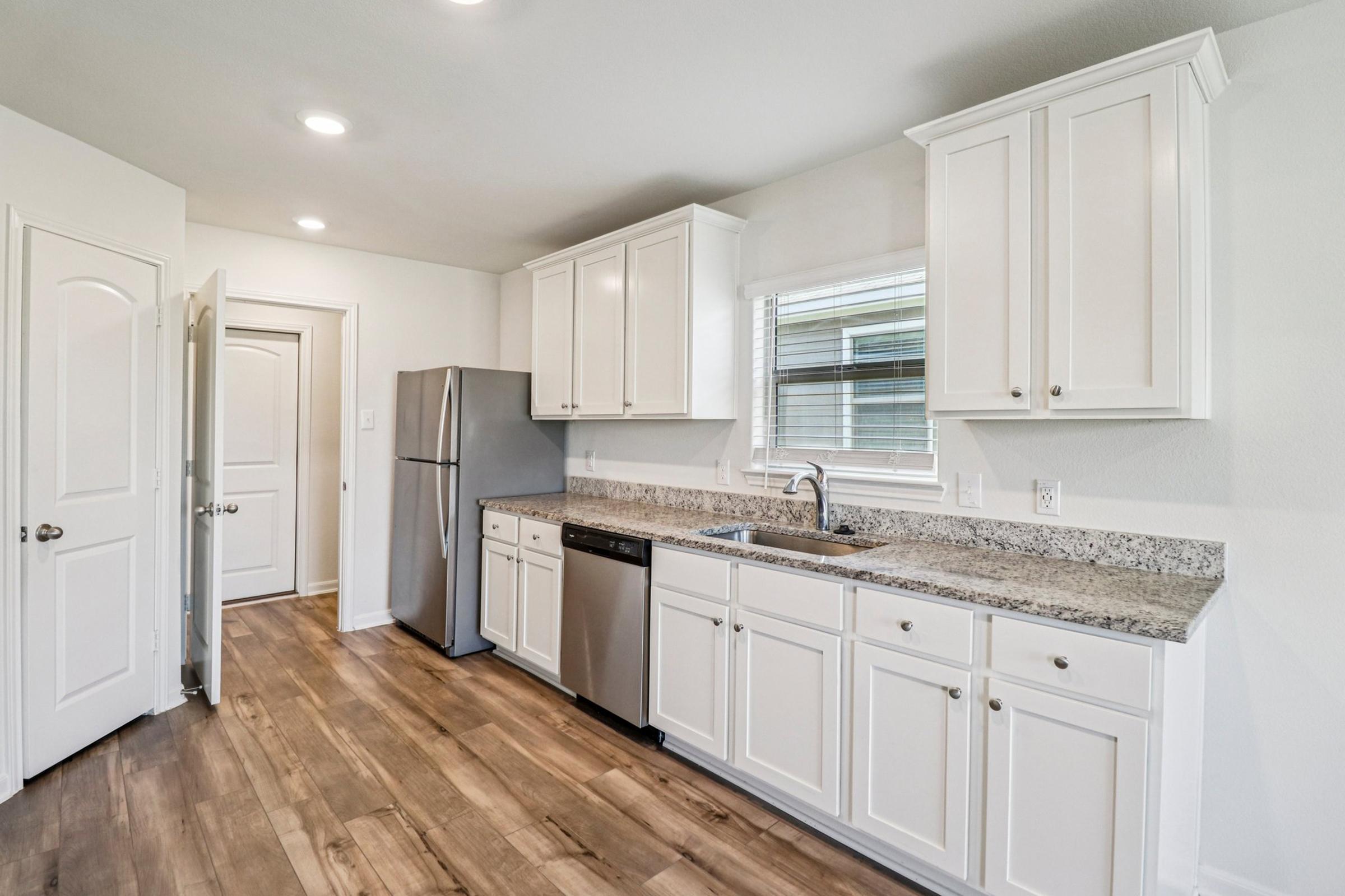 Modern kitchen featuring white cabinetry, stainless steel appliances, and a granite countertop. The space has an open layout with hardwood flooring and a window providing natural light. A refrigerator and dishwasher are included, and there's an adjacent doorway leading to another room.