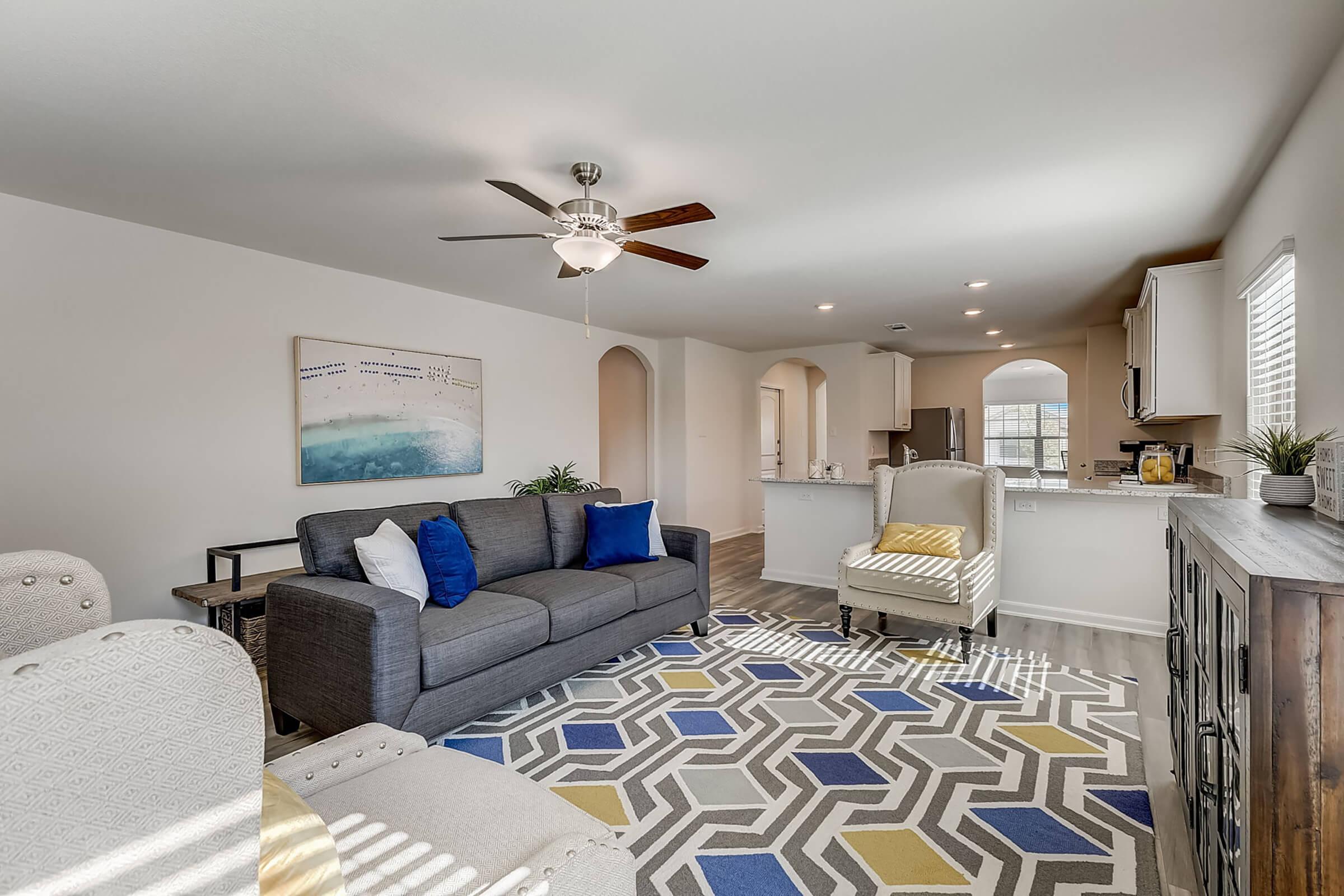 A modern living room featuring a gray sofa with blue and yellow pillows, a patterned area rug, and a ceiling fan. In the background, there is an open kitchen area with white cabinets and a dining space. Natural light streams in through the windows, creating a bright and inviting atmosphere.