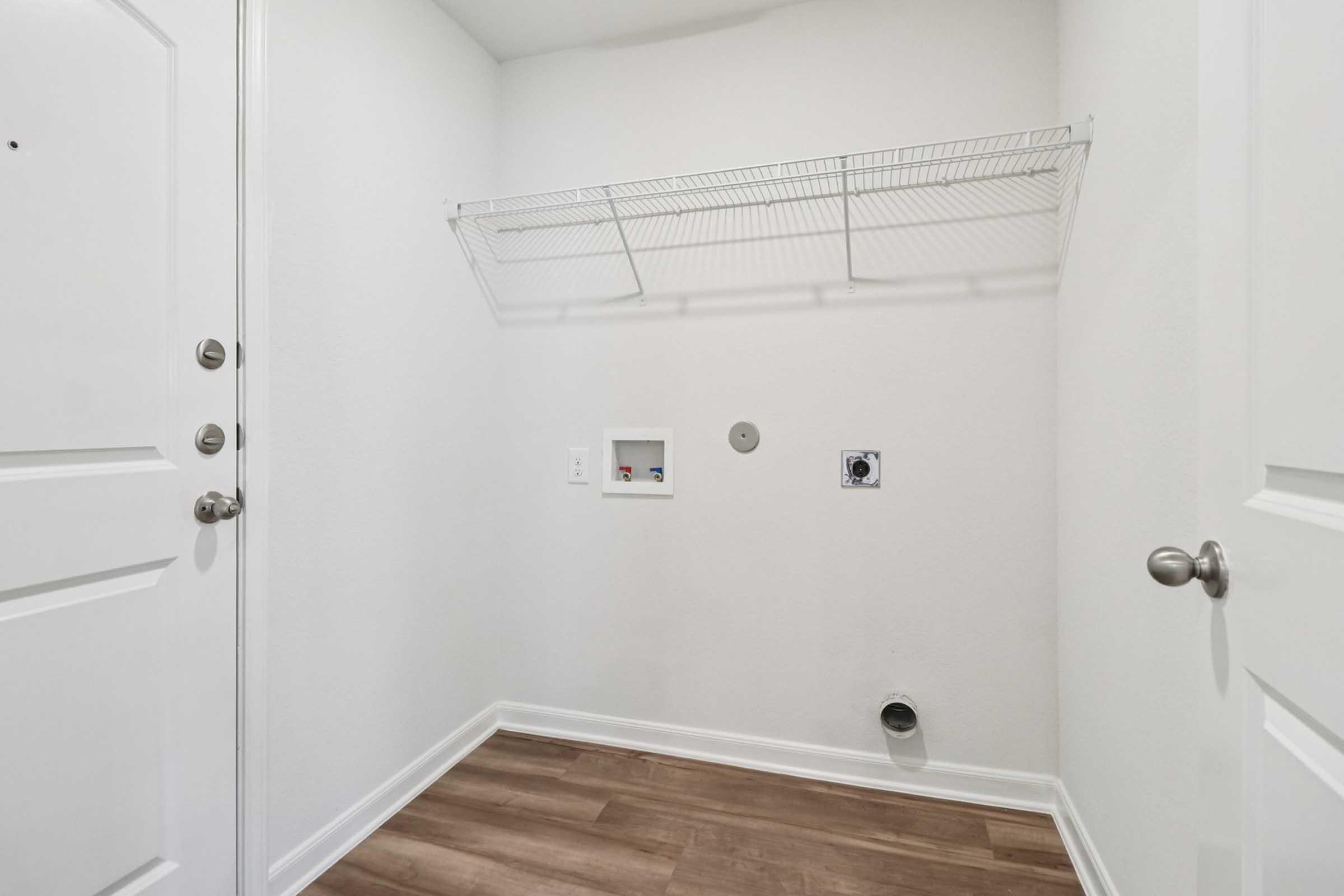 A clean, empty laundry room with white walls and a wooden floor. A shelf is mounted on the wall for storage, and there are utility connections and an outlet visible. The door to the room is closed, and the entrance is equipped with two metal handles.