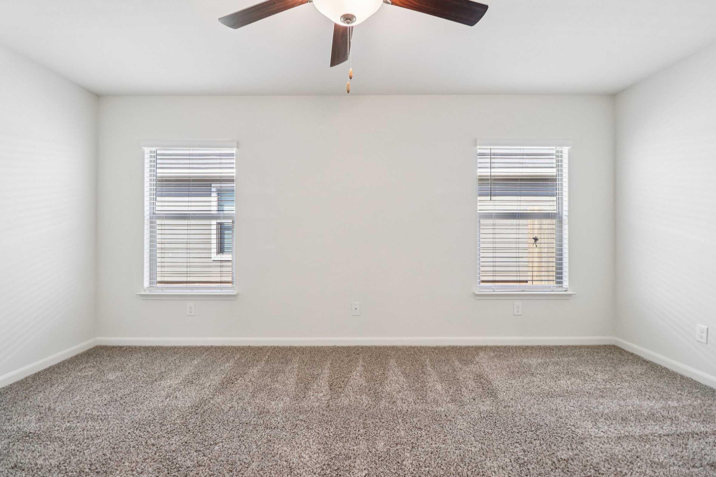 A bright, empty room featuring light-colored walls and two windows with blinds. The space has a ceiling fan and a plush carpeted floor, creating a cozy atmosphere. Natural light streams in through the windows, enhancing the airy feel of the room.