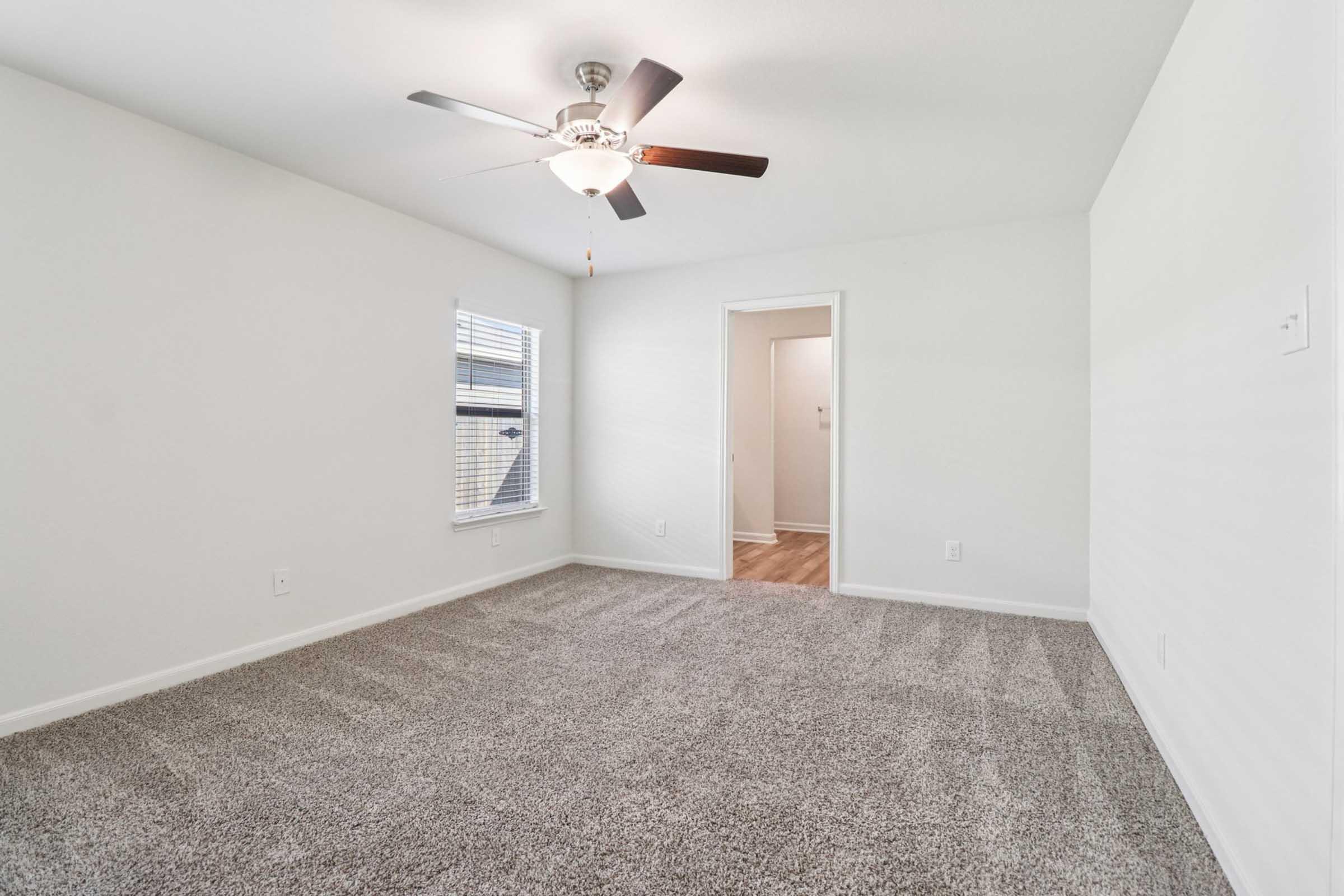 A spacious, empty room featuring light-colored walls and a plush carpet. There is a ceiling fan with a light fixture, a window allowing natural light, and an open doorway leading to another room, creating a bright and inviting space.