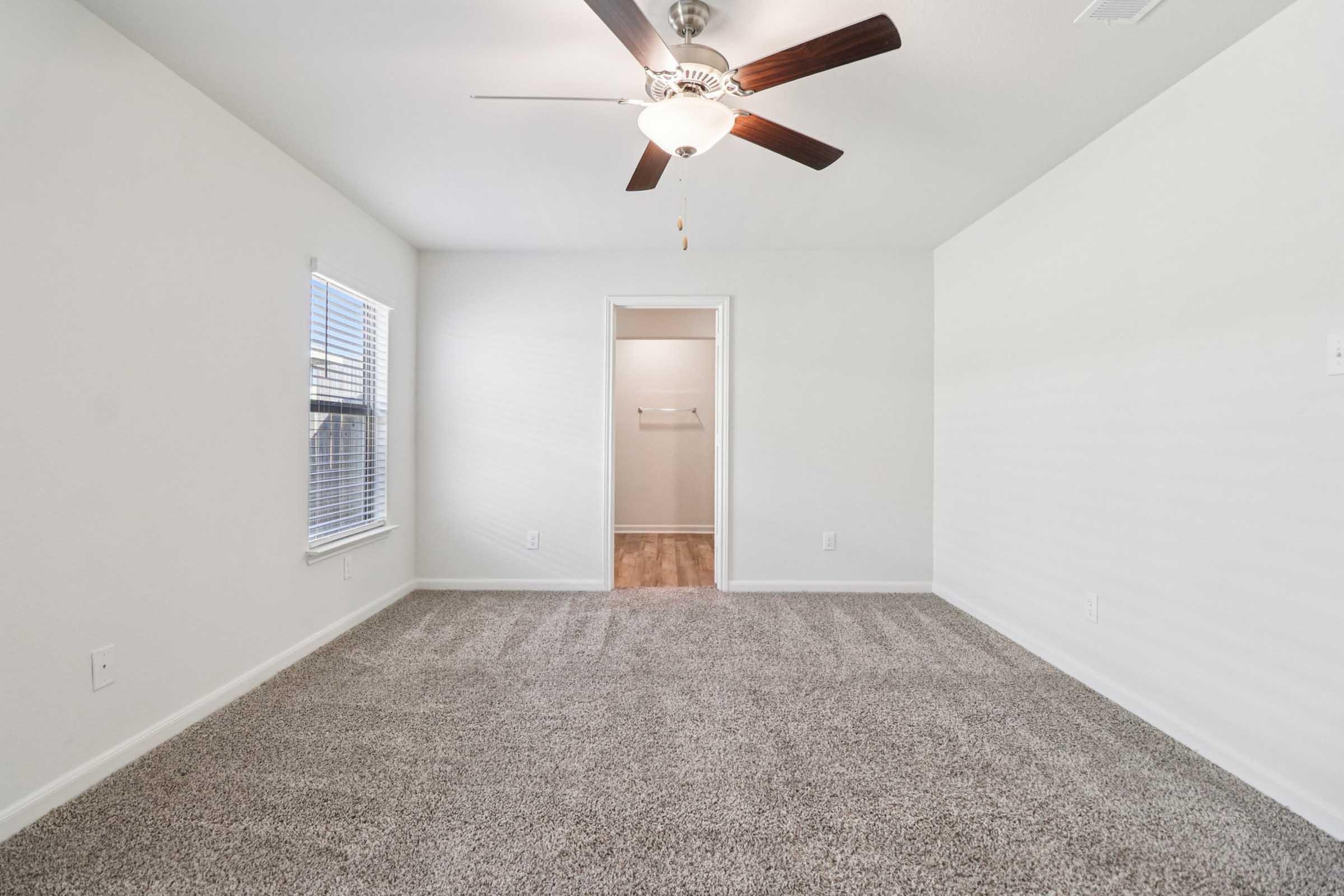 A bright, empty bedroom featuring light-colored walls, a ceiling fan, and a large window with blinds. The spacious room has plush beige carpeting and a doorway leading to a closet with a hanging rod visible. The overall atmosphere is clean and inviting, suitable for personal decoration.