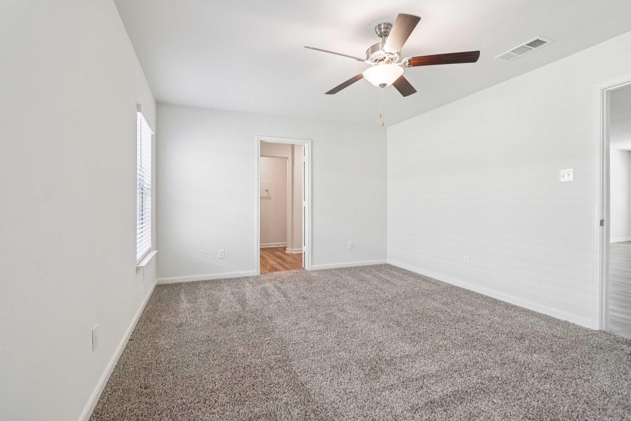 A spacious, empty room featuring light-colored walls and a ceiling fan. The floor is covered in plush gray carpet. A doorway leads to a small room or closet, with a large window allowing natural light to fill the space. The overall atmosphere is bright and inviting, suitable for various furniture arrangements.