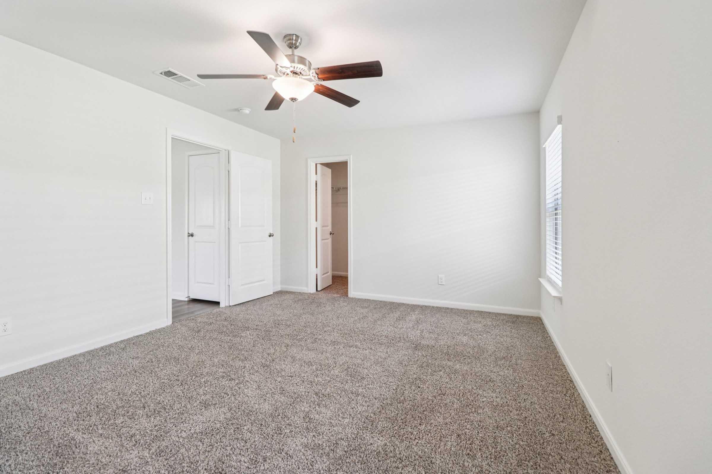 An empty room featuring light-colored walls and a plush carpet. A ceiling fan is mounted in the center, and there are two doors leading to other areas. Natural light enters through a window on the far wall, creating a bright and airy atmosphere.