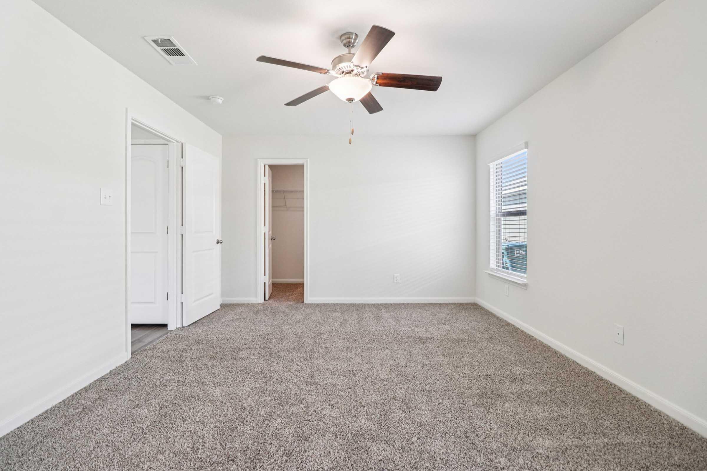 A well-lit, empty room featuring light gray carpet, a ceiling fan with wooden blades, and white walls. There are two doors—one leading to a closet and the other to a hallway. A window on the right provides natural light, enhancing the spacious and clean appearance of the room.