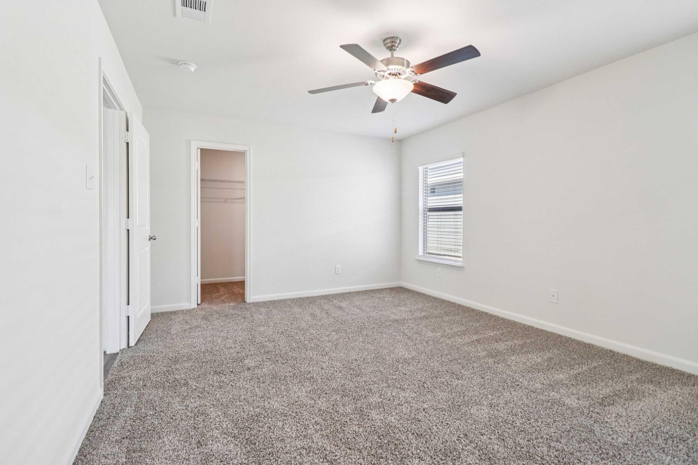A bright and spacious bedroom with light-colored walls and carpeted flooring. It features a ceiling fan, a window with blinds, and an open doorway leading to a closet area. Natural light fills the room, creating a welcoming atmosphere.