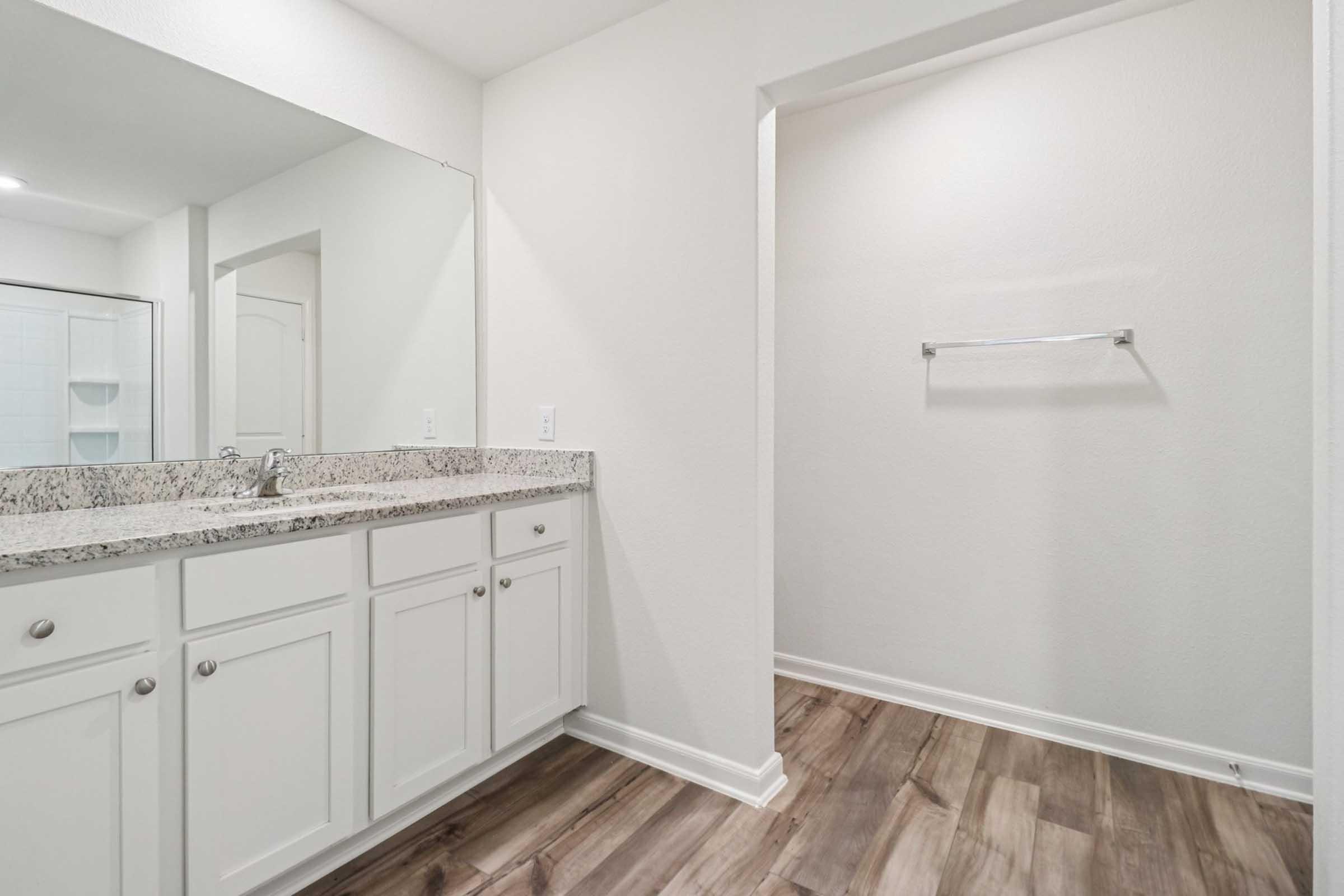 A modern bathroom featuring a granite countertop with a sink and white cabinet below. A large mirror reflects the space, and to the right, there is an open area with a towel rack. The floor is made of light-colored wood, and the walls are painted in neutral tones, creating a clean and spacious look.