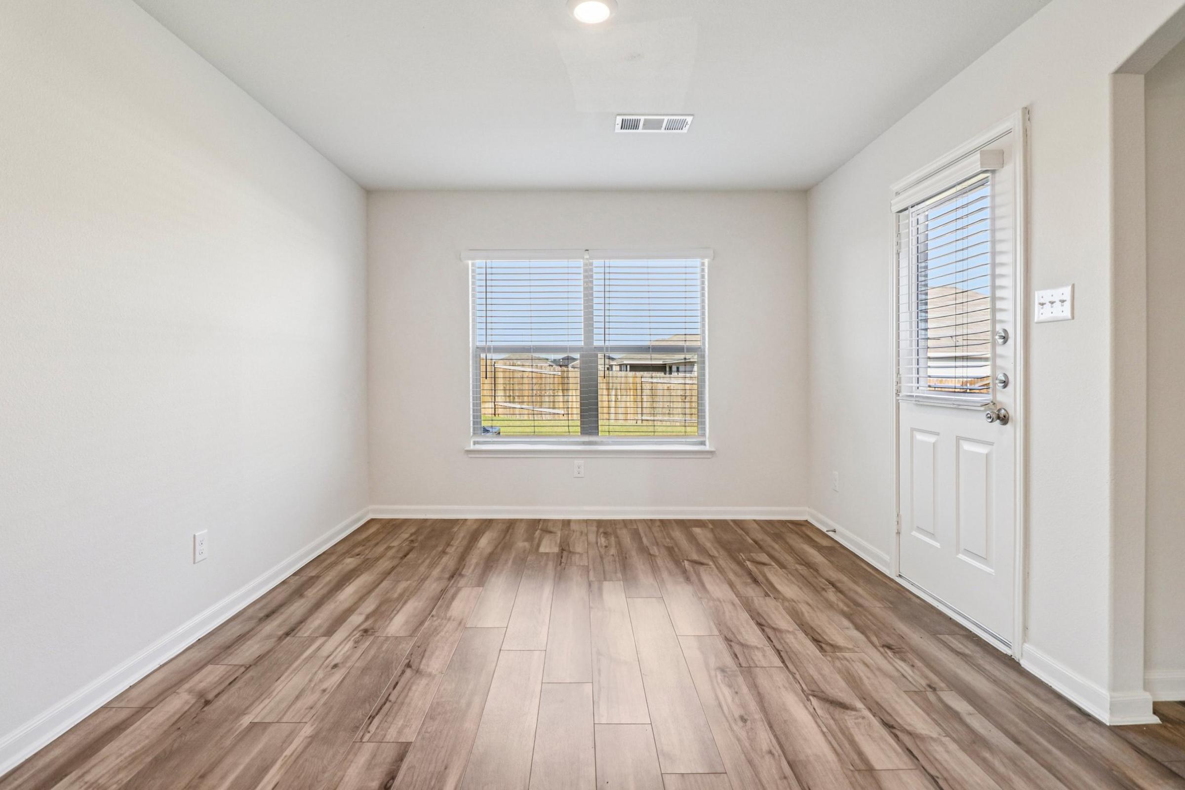 Bright, empty room with light-colored walls and wooden flooring. A large window provides natural light, and there is a door leading outside. The space appears clean and modern, suitable for various purposes such as a living area or office.