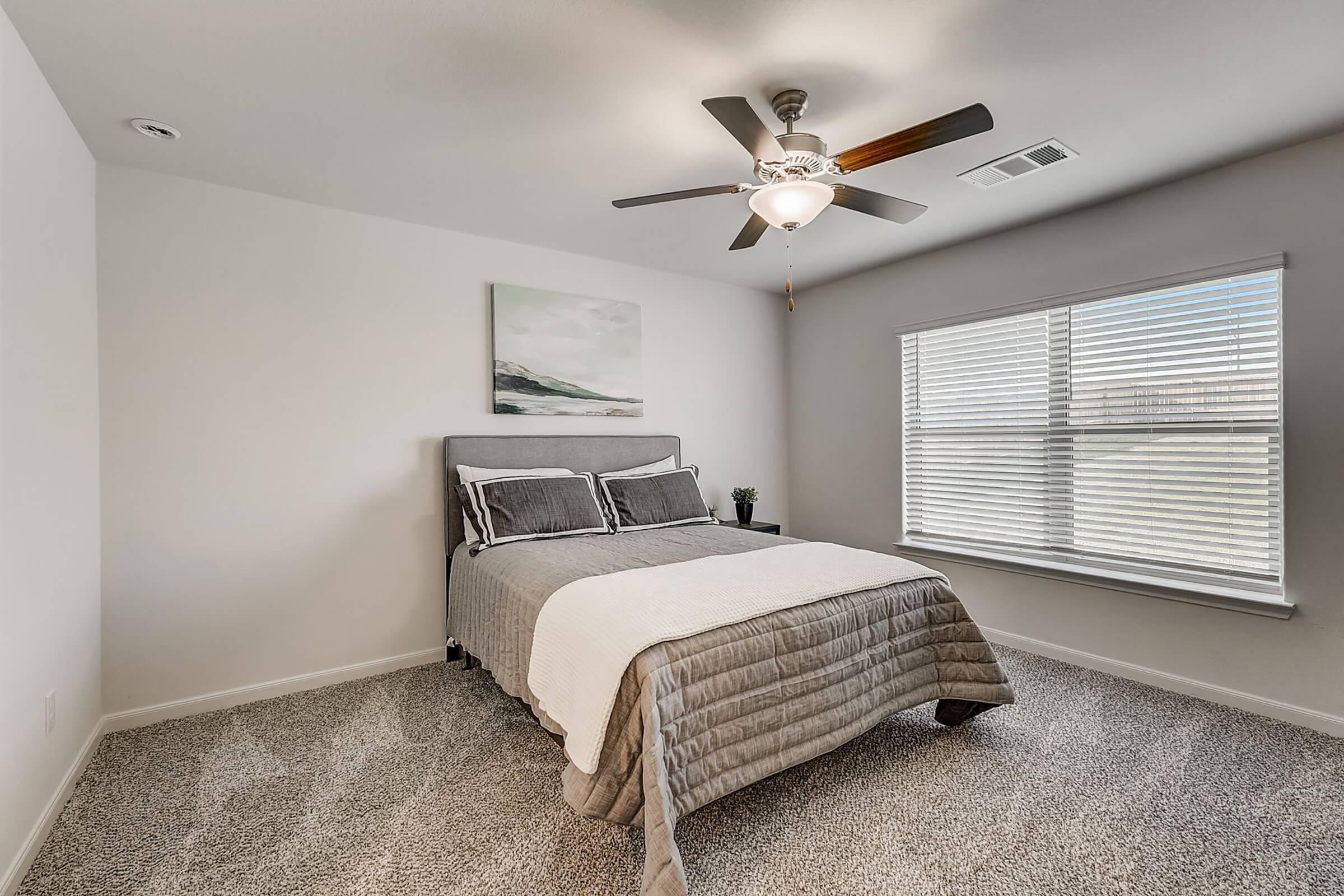 A cozy bedroom featuring a neatly made bed with a light gray quilt, two decorative pillows, and a ceiling fan overhead. Natural light streams through a window adorned with blinds, while a simple landscape painting hangs on the wall. The room has a warm, inviting atmosphere with soft carpeting.