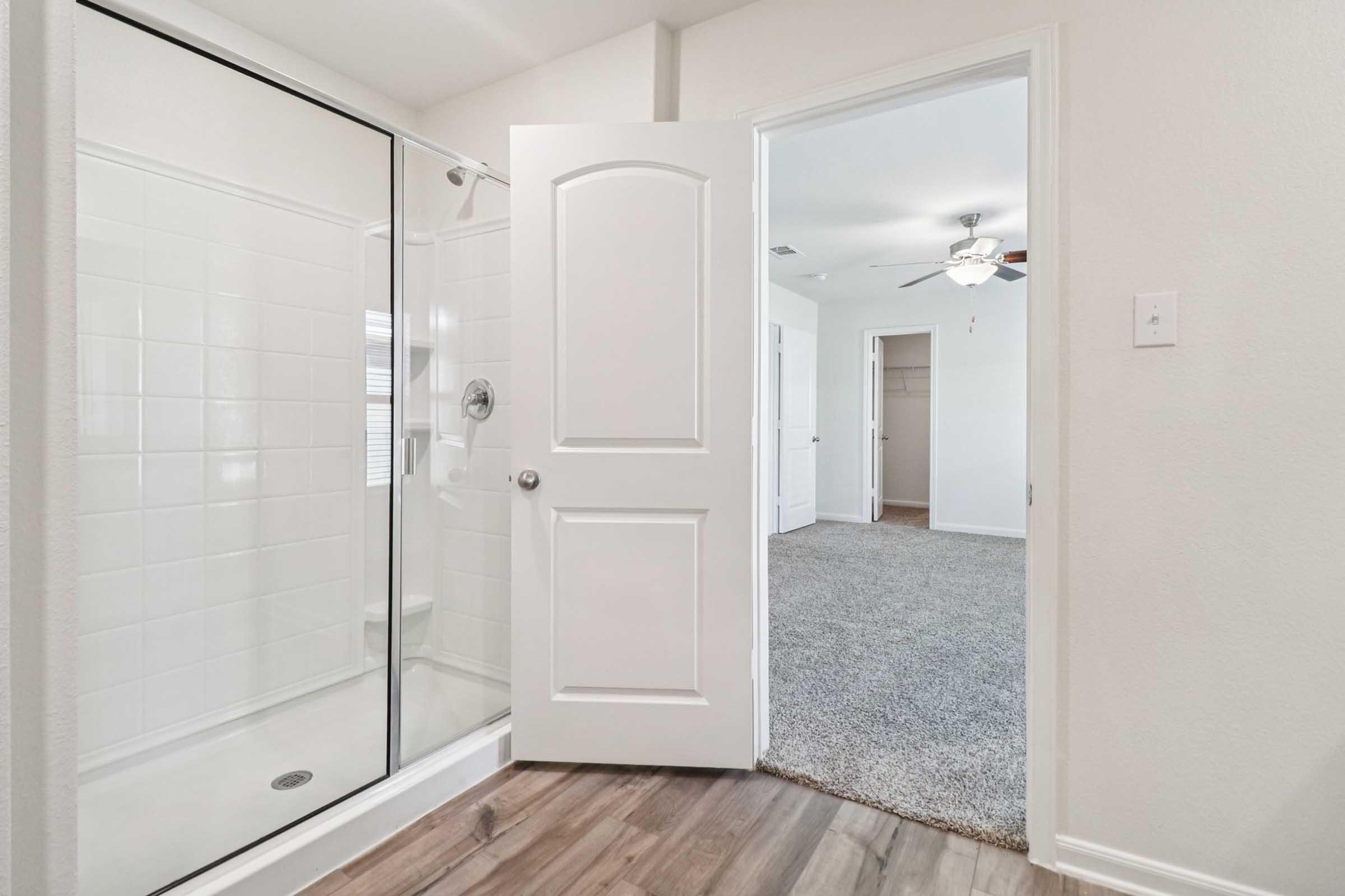 Interior view of a bathroom featuring a glass-enclosed shower on the left and an open door leading to a carpeted bedroom on the right. The walls are painted light colors, and there is a ceiling fan visible in the adjacent room. The flooring consists of wood-like laminate.
