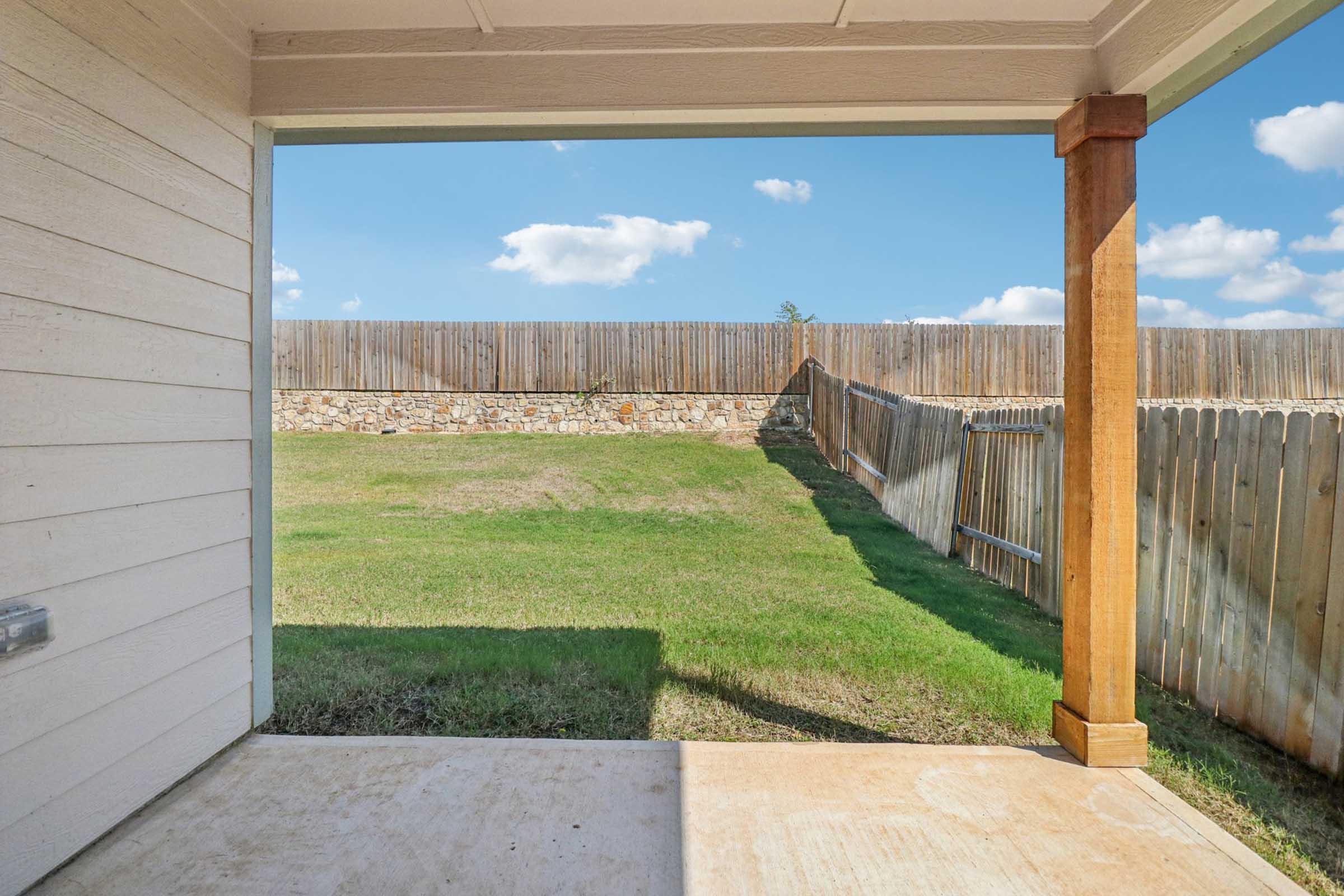 View from a covered patio looking out onto a grassy backyard with a wooden fence. The sky is bright blue with a few fluffy clouds. The ground is mostly clear concrete, and the area is well-lit by sunlight. A stone wall is visible along one side of the yard, providing a natural boundary.