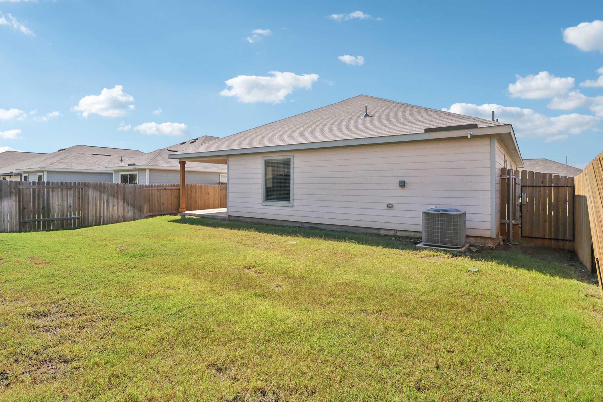A view of the exterior of a single-story house featuring a fenced backyard. The house has a light-colored siding, a shaded patio area, and a visible air conditioning unit. The lawn is well-maintained with green grass, and the sky is partly cloudy.