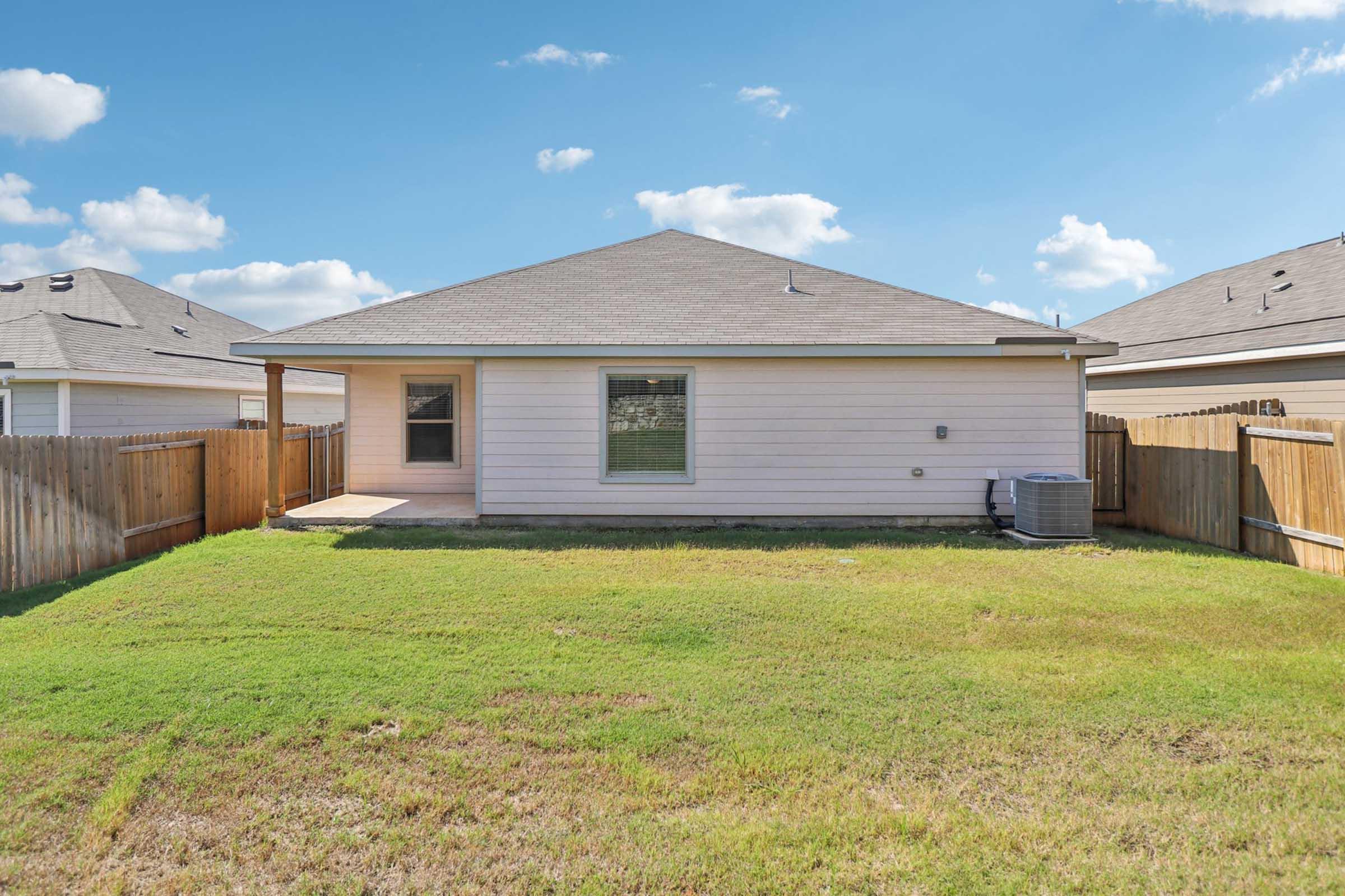 A view of the backyard of a single-story house, featuring a grassy yard enclosed by wooden fencing. The house has a small covered porch and a window facing the yard, with an air conditioning unit visible on the side. Clear blue skies and scattered clouds are in the background.
