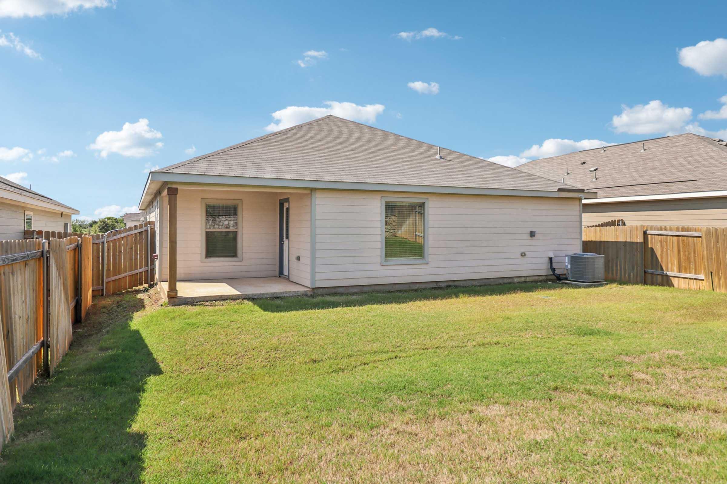 Backyard view of a single-story house with a light-colored exterior, featuring a small patio area and a fenced yard. The lawn is well-maintained with green grass, and the sky is bright with a few clouds. An air conditioning unit is visible beside the house.