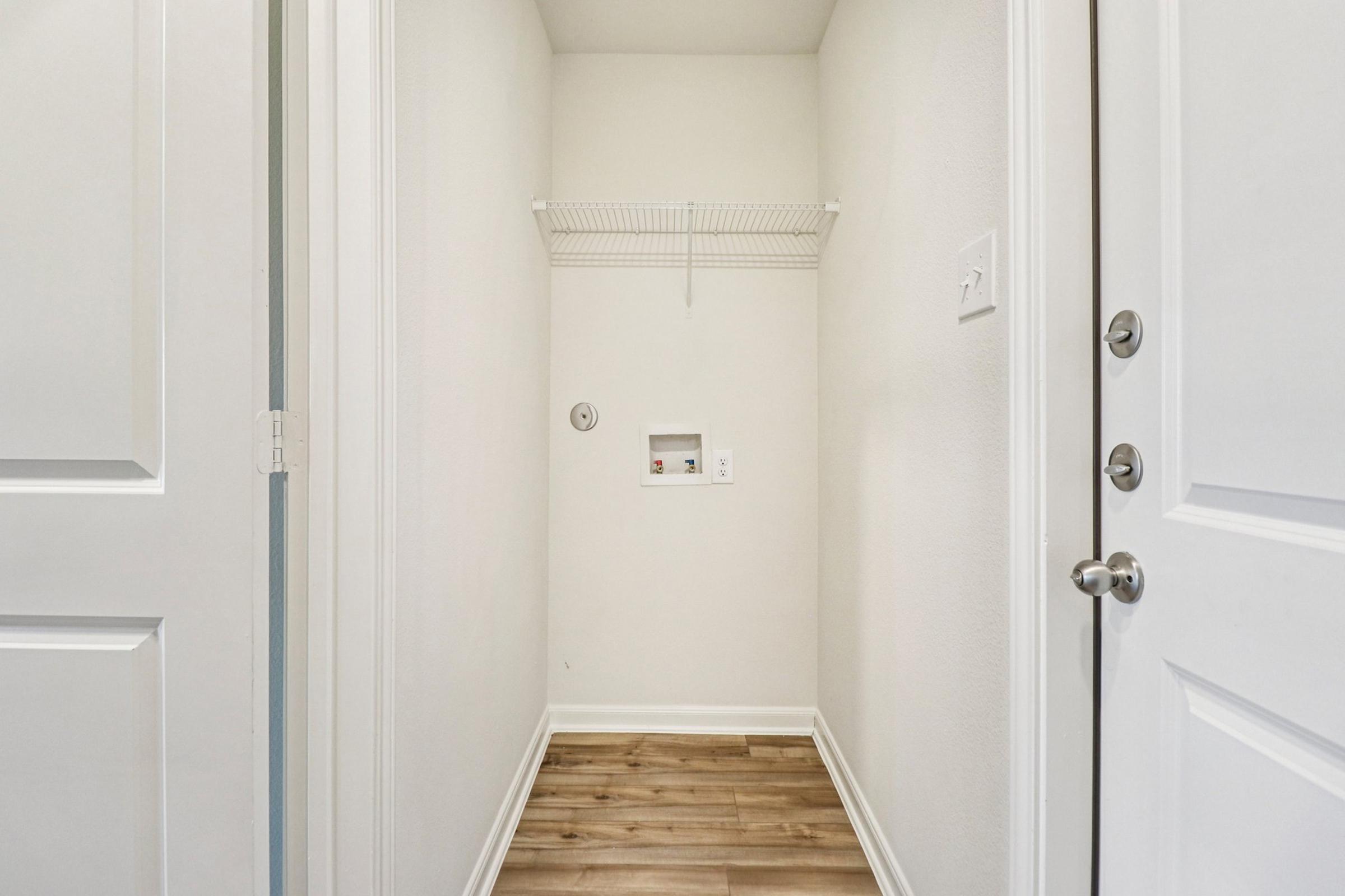 A small, empty laundry area inside a home. The walls are painted white, and there is a wire shelf on the left side. A door is visible on the right side, leading outside. The floor is made of light-colored laminate wood. There is a power outlet and a small utility box on the wall.