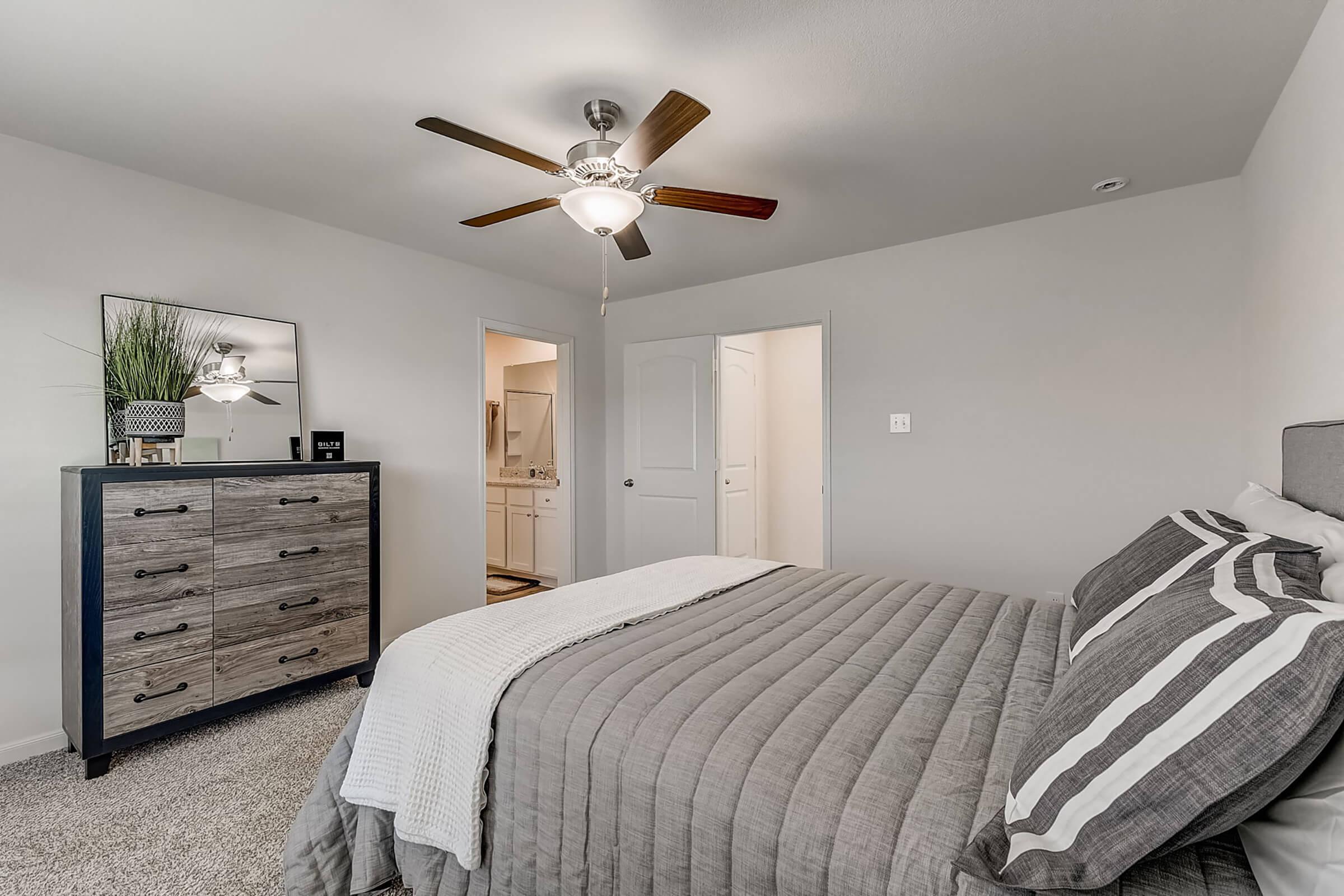 A stylish bedroom featuring a gray-themed decor. The room includes a neatly made bed with striped pillows, a ceiling fan, a dresser with a mirror, and an open doorway leading to a bathroom. The soft carpet and neutral walls create a cozy atmosphere.
