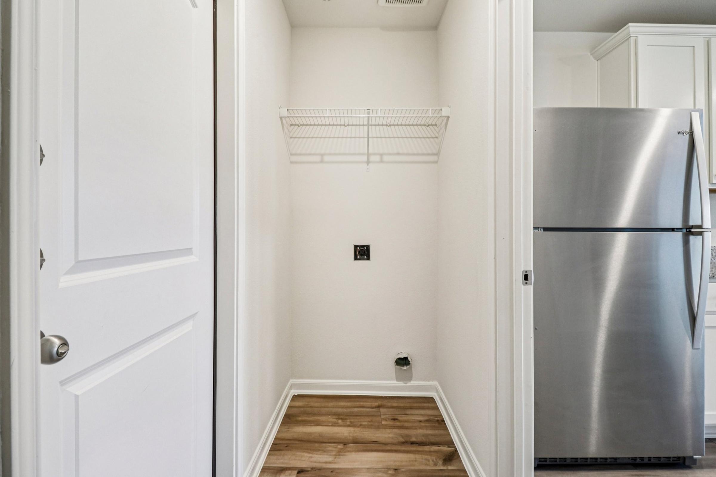A small, empty laundry room with white walls and a wooden floor. A wire shelf is mounted on the wall, and there is a power outlet visible. The corner shows a vent or pipe, and there is a stainless steel refrigerator nearby, partially visible on the right side.
