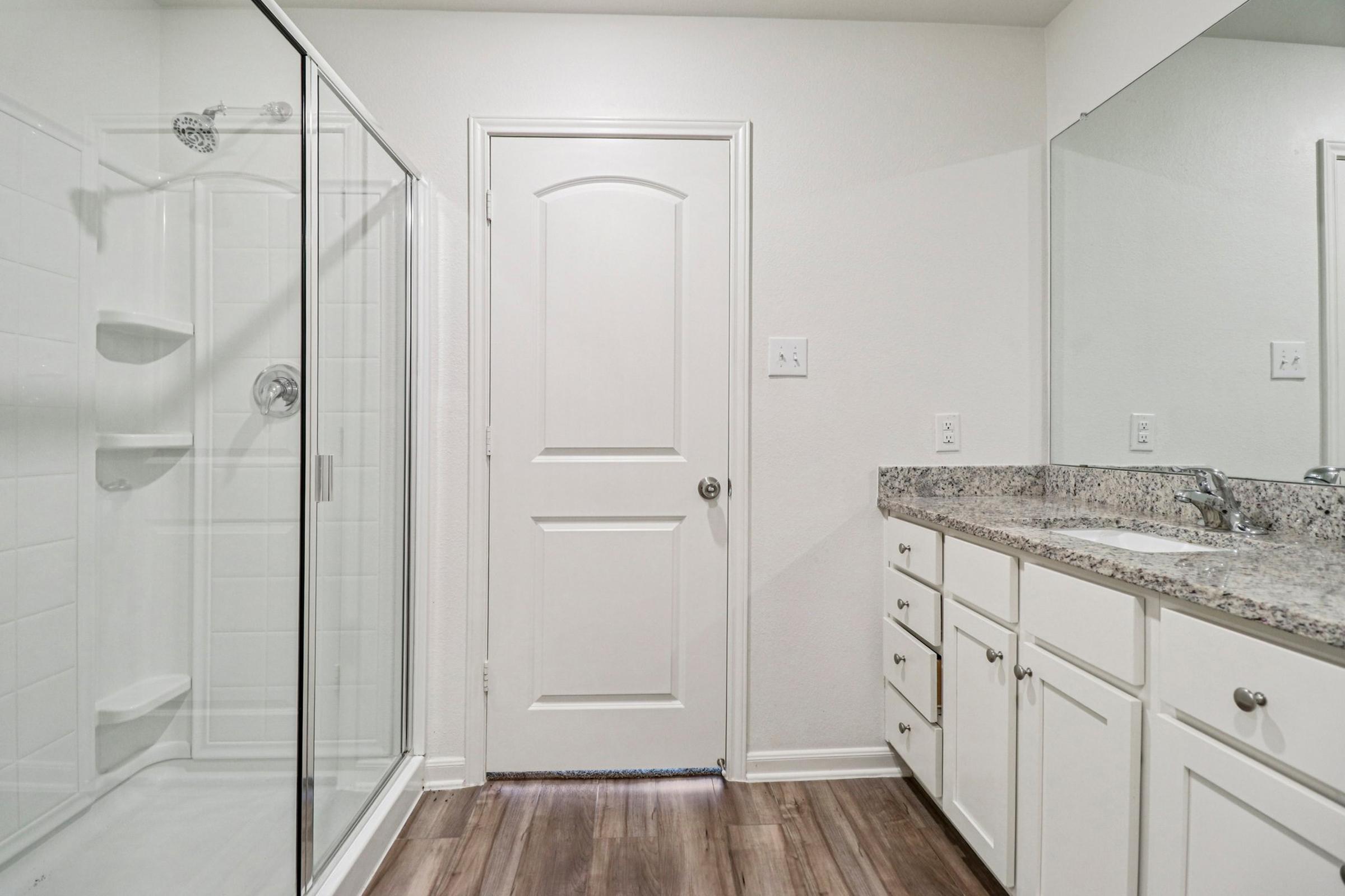A modern bathroom featuring a glass-enclosed shower, a single sink vanity with granite countertop, and a large mirror. The walls are painted in a light color, and there is a wooden floor. A white door is visible on the right, leading to another room. The space is well-lit and clean.