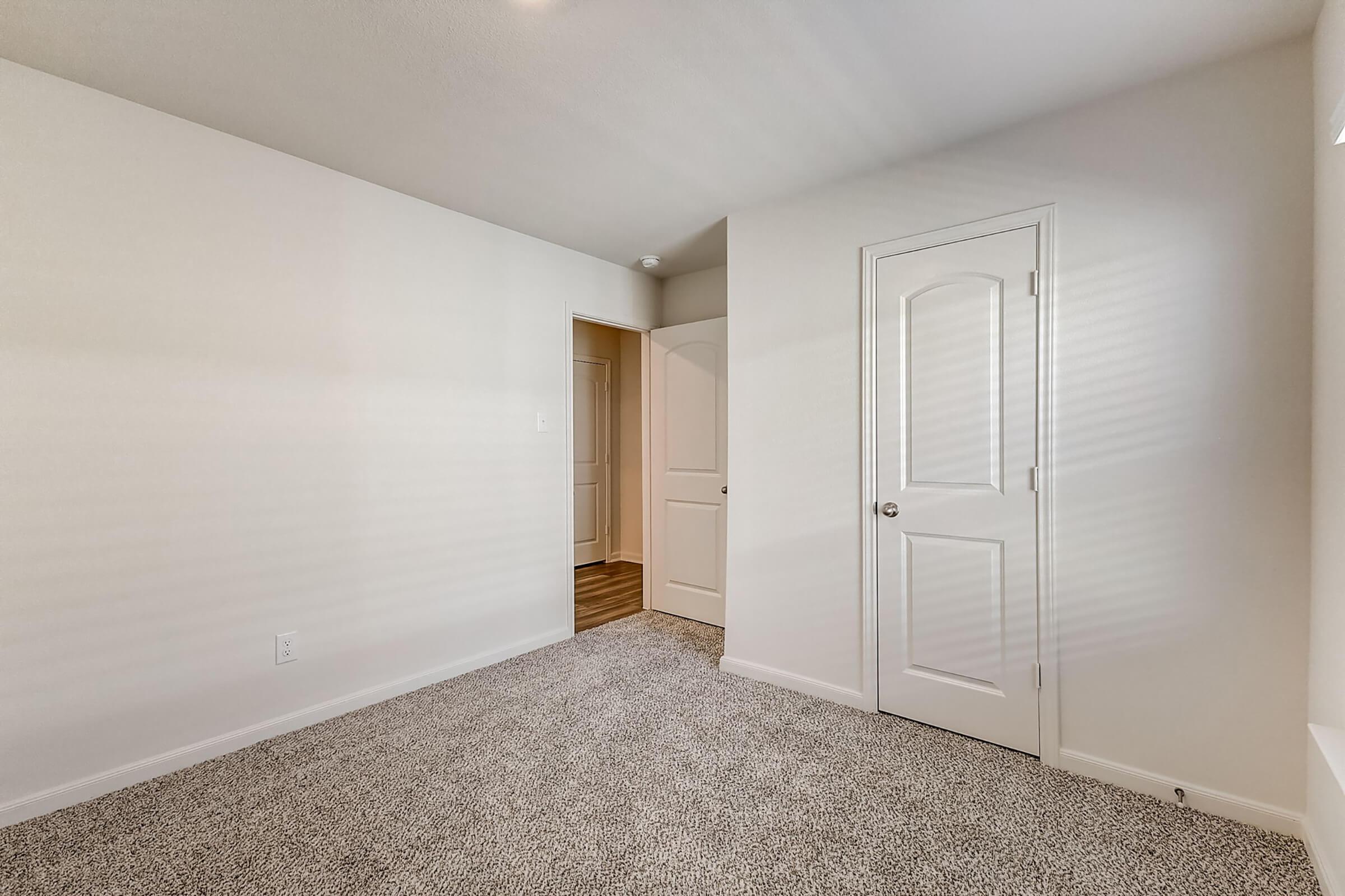 A clean, minimalist room featuring beige carpet flooring, light-colored walls, and a closed door leading to another space. Natural light creates soft shadows, enhancing the simple decor. The room has no furniture, providing an open and airy feel ideal for personalization.