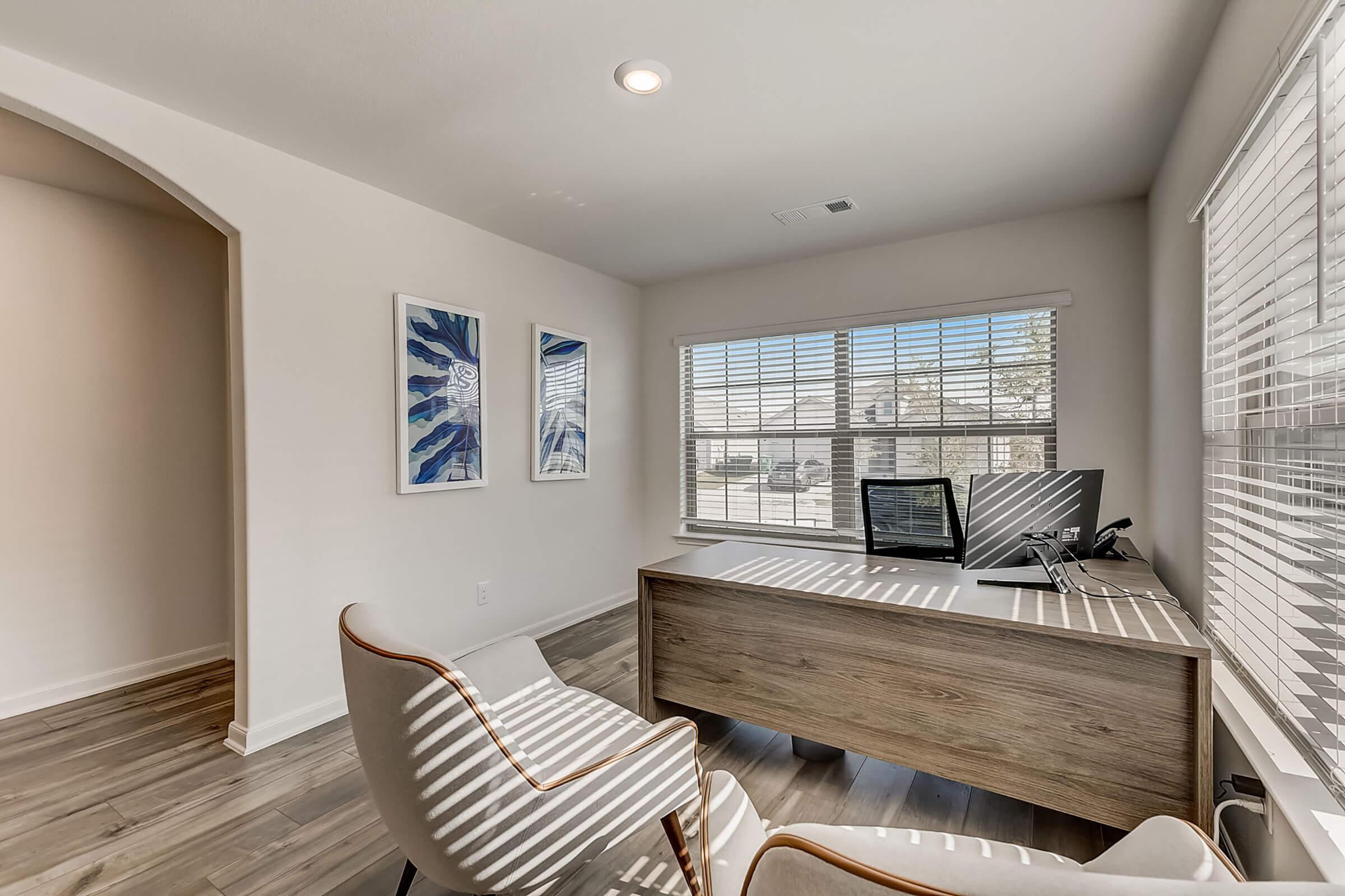 A modern home office with a wooden desk, a computer setup, and an ergonomic chair. The room features large windows with blinds allowing natural light to enter, illuminating the space. On the walls, there are two framed artworks, and light wooden flooring complements the airy atmosphere.