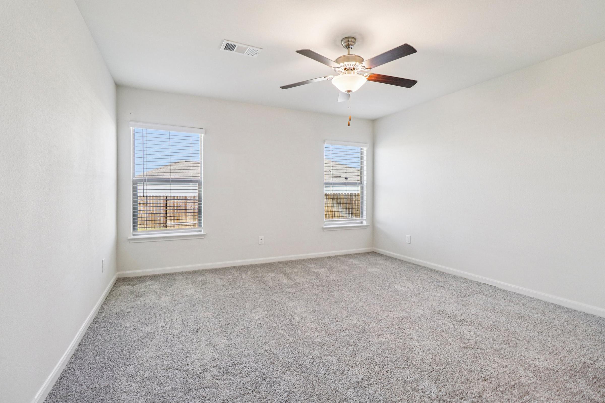 A spacious, empty room with light-colored walls and a ceiling fan. The floor is covered in soft gray carpet. Two windows provide natural light, showcasing a clear view outside. The room is well-lit and ideal for various uses, such as a bedroom or office.