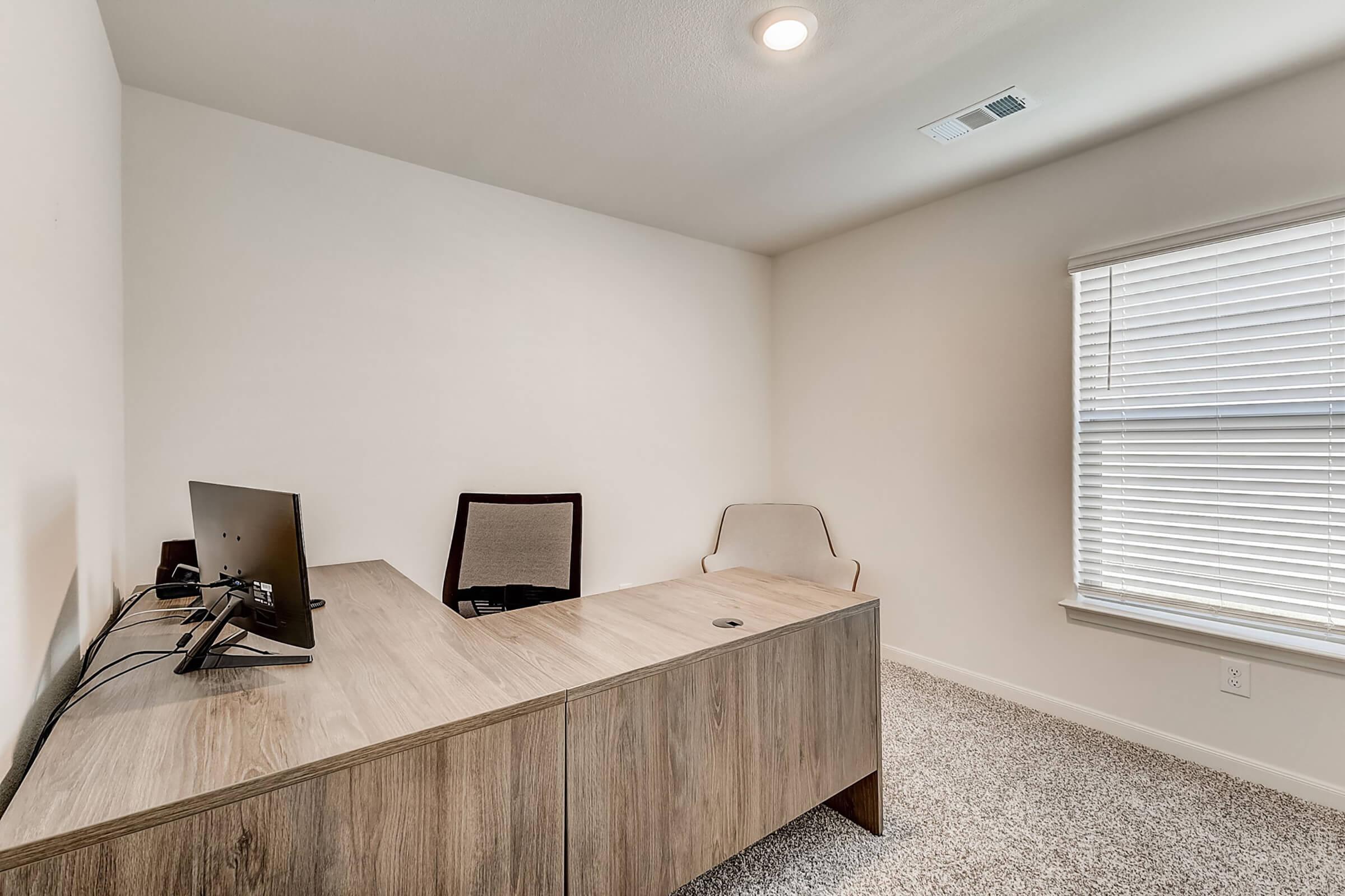 A modern, minimalistic office space featuring a wooden desk with a computer monitor, an ergonomic chair, and a second chair. The walls are painted white, and there is a window with blinds, allowing natural light to brighten the room. The carpeted floor adds warmth to the interior.