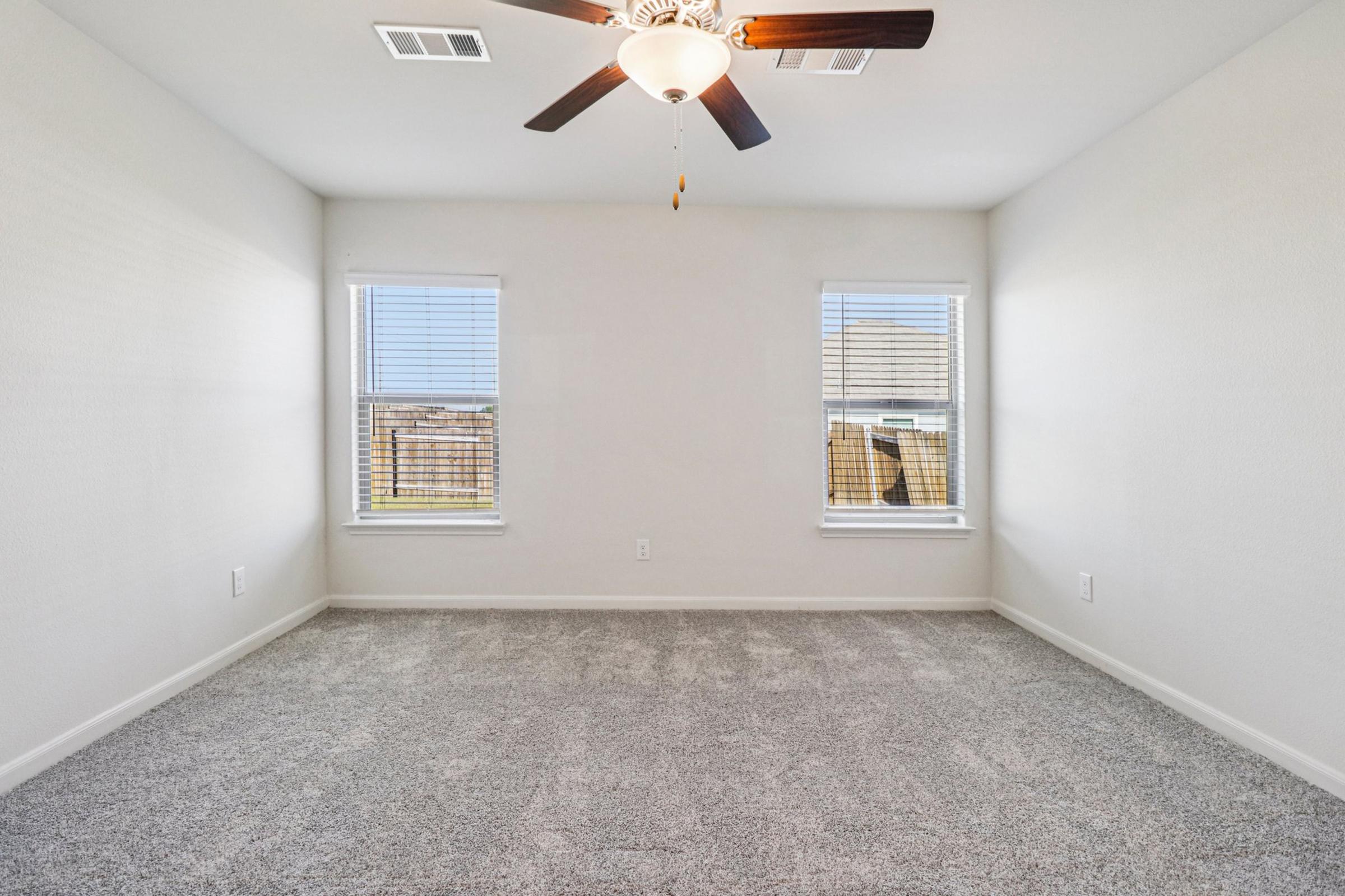 A empty, well-lit room with gray carpet, featuring a ceiling fan and two windows with blinds. The walls are painted white, and there are no furniture or decorations. Natural light streams in from the windows, creating a bright and inviting atmosphere.