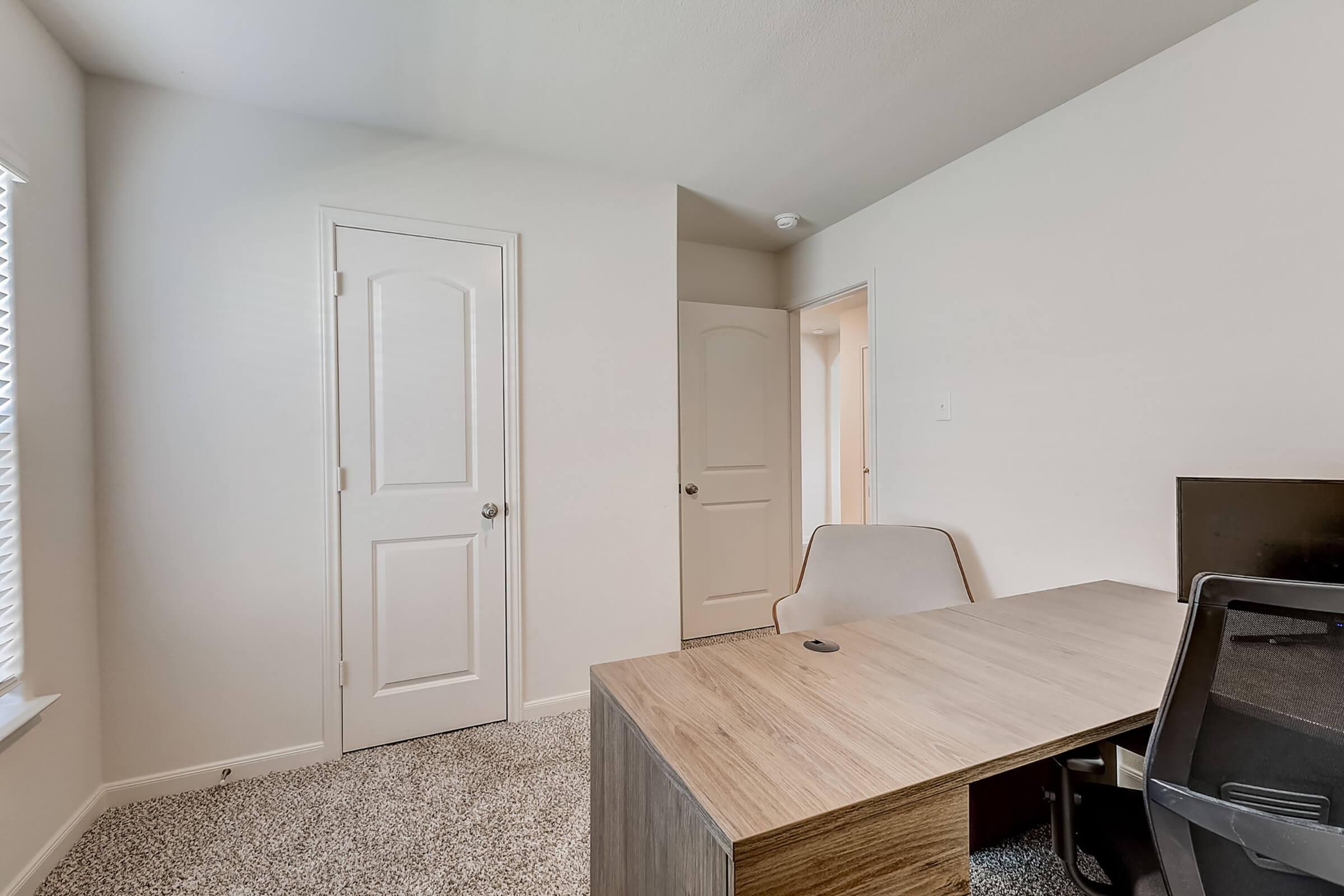 A light-colored room featuring a simple wooden desk and a chair. Two doors are visible, one leading to another area, with ample natural light coming from a window. The flooring is carpeted, and the walls are painted in a neutral tone, creating a clean and modern workspace atmosphere.
