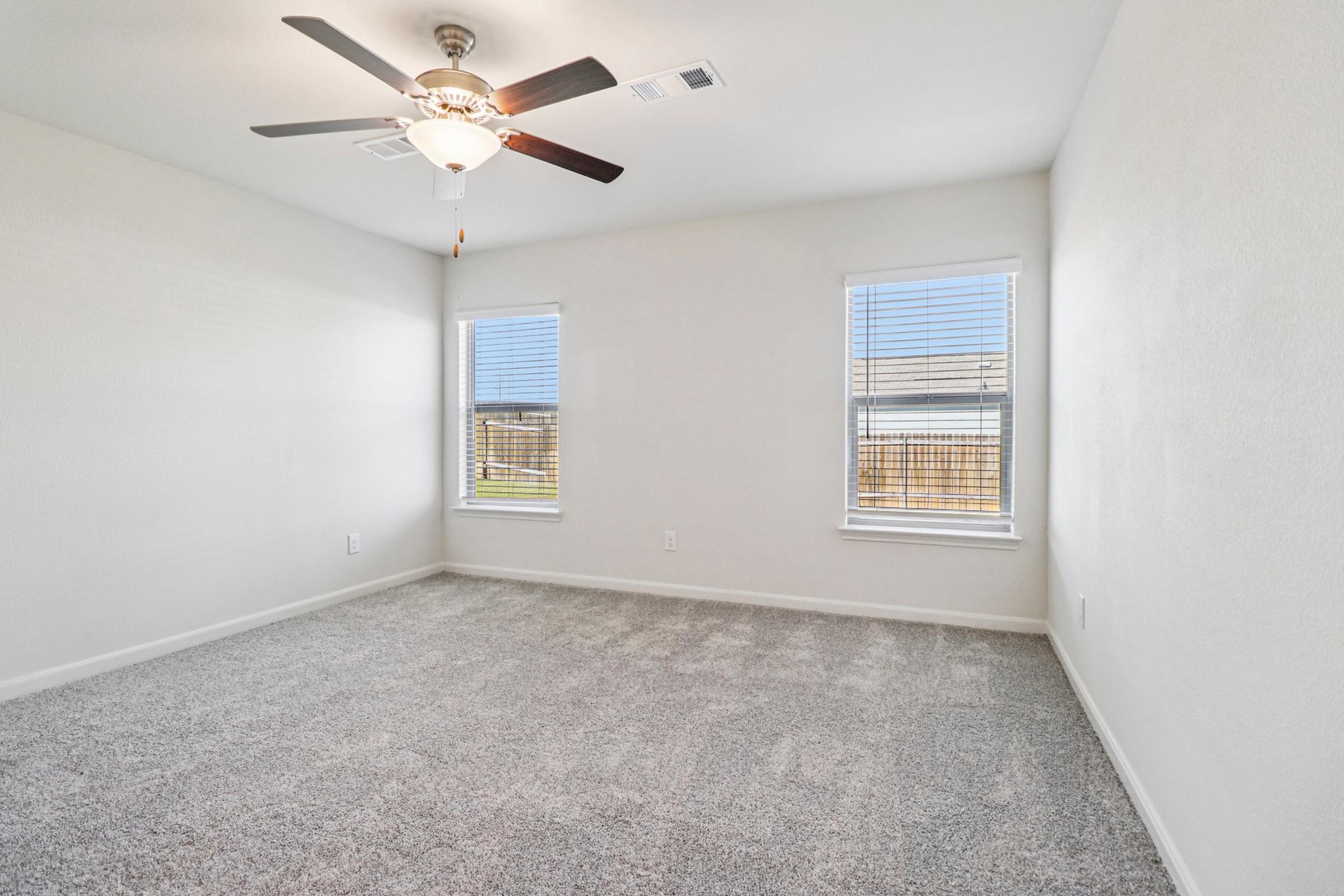 Empty room with light-colored walls and a plush carpet. A ceiling fan is mounted in the center, and there are two windows on opposite walls allowing natural light to enter. The overall space appears bright, clean, and ready for furnishing.