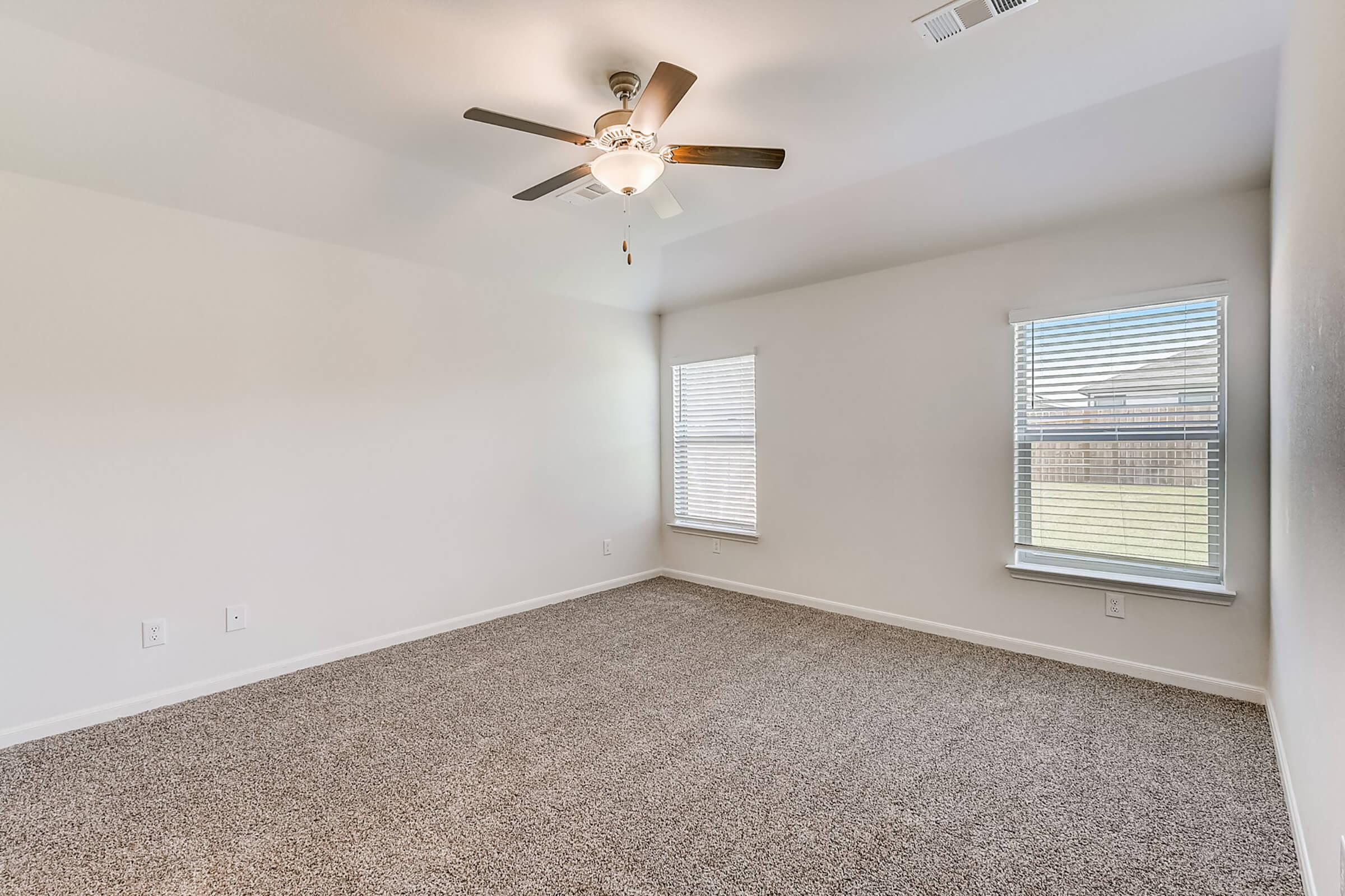 A bright, empty room with light beige carpet, two windows with white blinds, and a ceiling fan. The walls are painted white, and the space has a clean, open feel, suitable for various decor styles. Natural light streams in through the windows, enhancing the airy atmosphere.