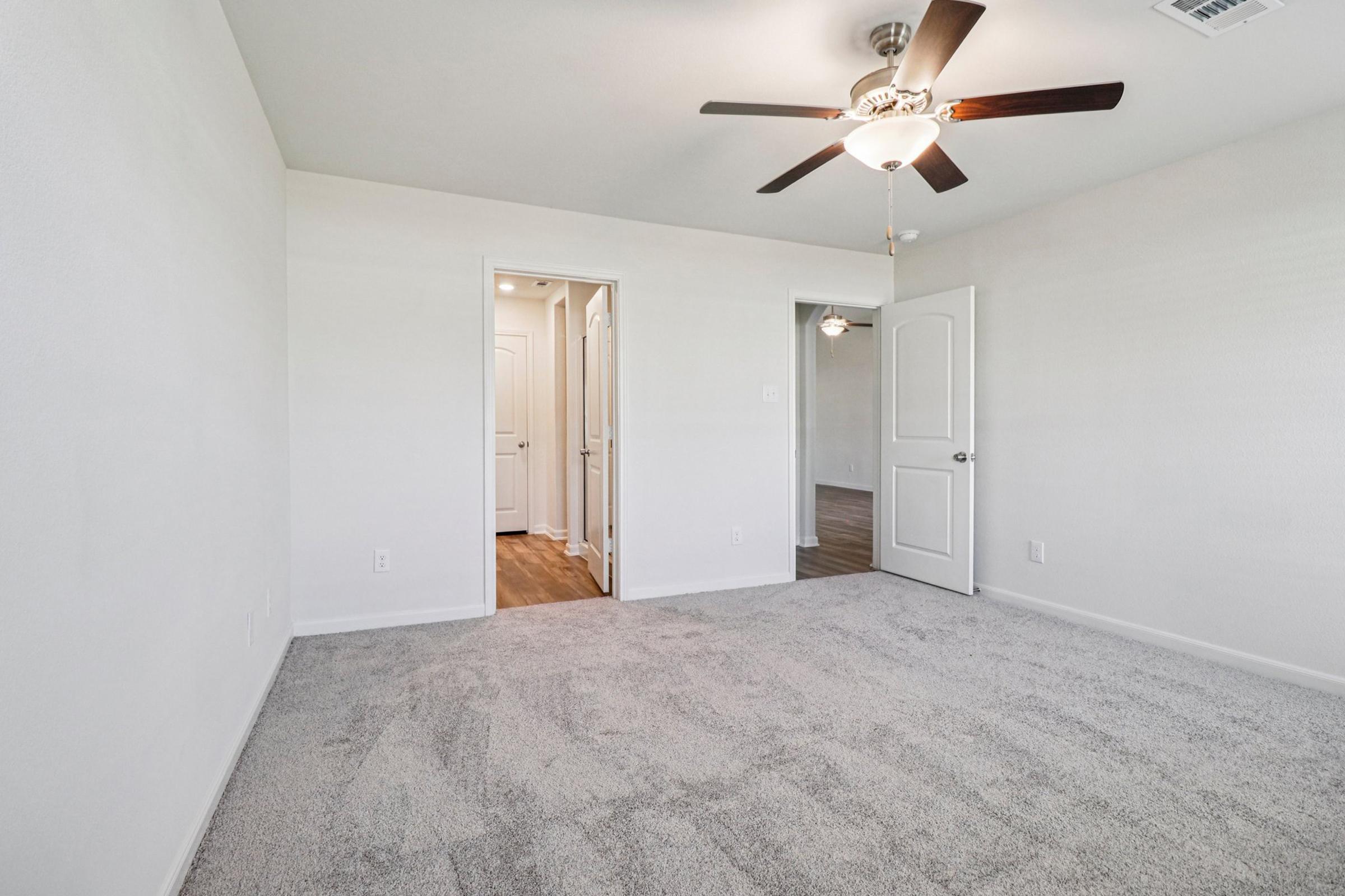 Empty room with light-colored walls and a plush carpet. A ceiling fan is installed, and there are two doorways leading to other areas. Natural light appears to enter from the hallway. The room is spacious and well-lit, creating an inviting atmosphere.
