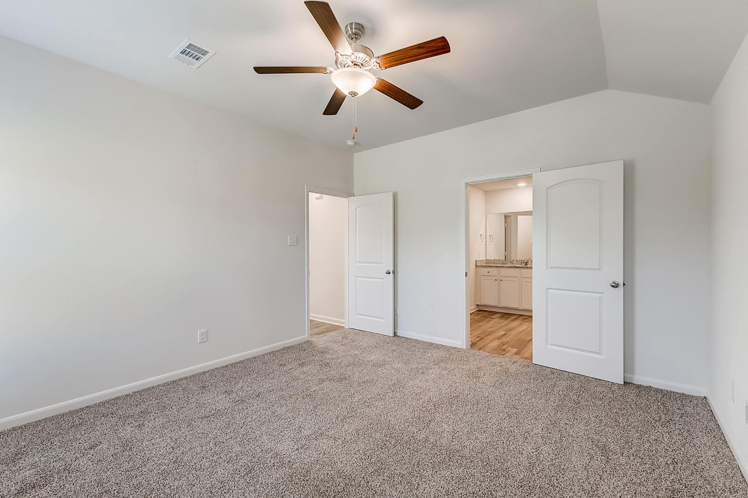 A well-lit, empty room with light gray walls and plush carpet. It features a ceiling fan and two doors leading to different areas, likely a closet and a bathroom. The room has a spacious feel, with natural light enhancing the open layout.