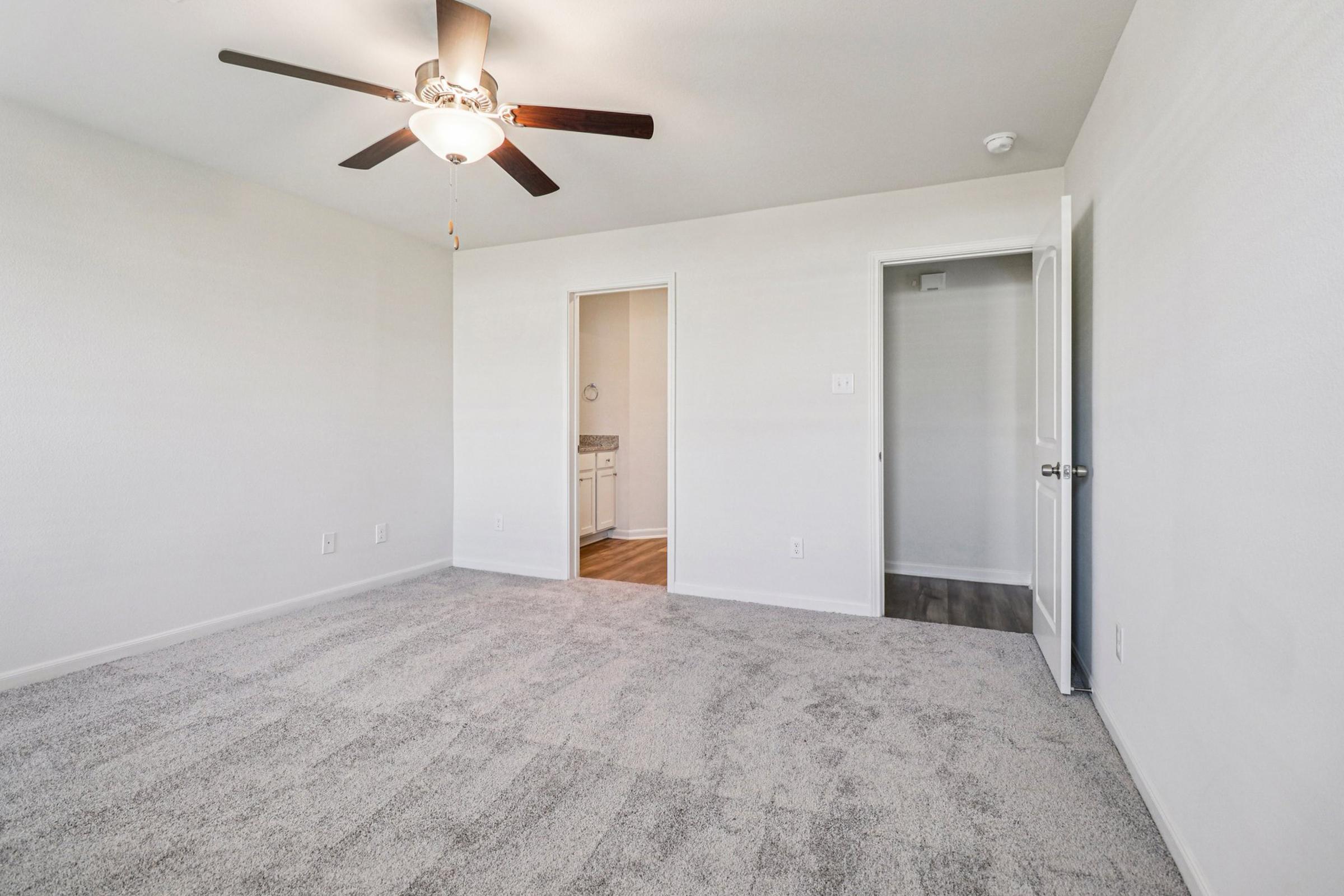A spacious, light-filled room with a ceiling fan and neutral-colored walls. The floor is covered with soft, gray carpet. Two doorways lead to other rooms, and a portion of the wall shows a small closet space. The overall atmosphere is bright and inviting, making it suitable for various uses.