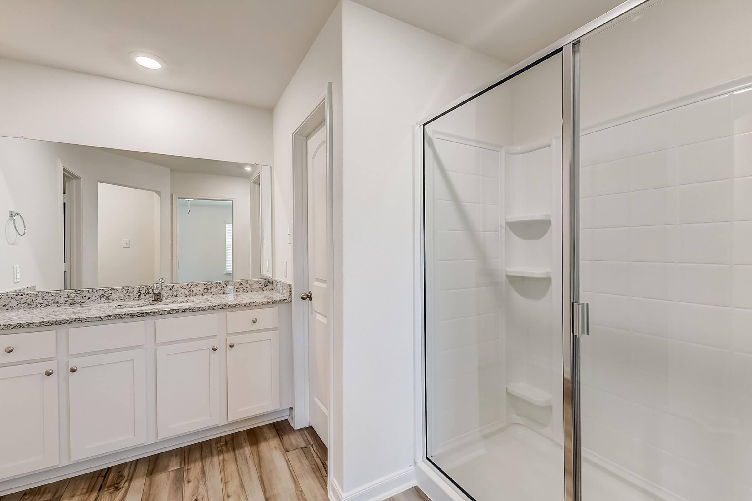 A modern bathroom featuring a glass-enclosed shower, white tiled walls, and a granite countertop with dual sinks. The space has light-colored walls and wooden flooring, with ample natural light from a nearby mirror reflecting the area.