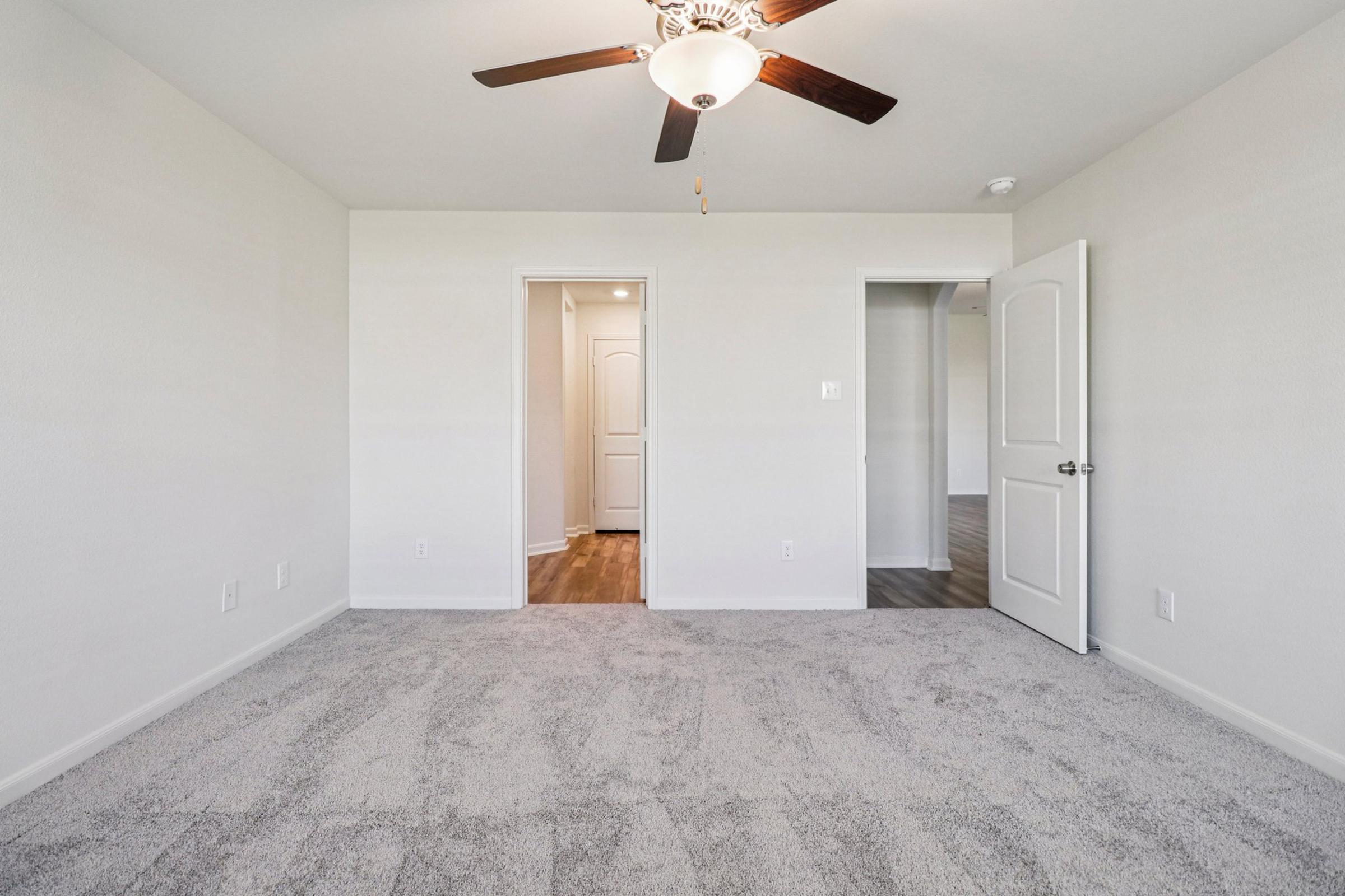 A spacious, empty room featuring light gray carpet and white walls. A ceiling fan is visible, and there are two doorways leading to other areas. Natural light is coming from an adjacent space, highlighting the clean, uncluttered atmosphere.