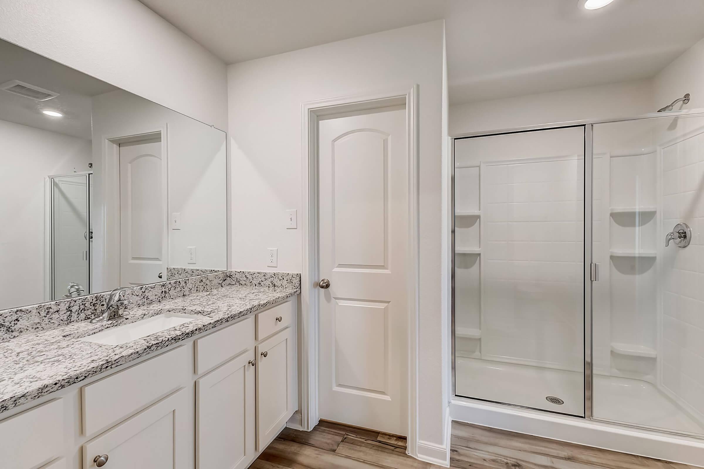 A modern bathroom featuring a double sink vanity with granite countertop, a large mirror, a closed door, and a glass-enclosed shower. The walls are painted white, and the flooring is a light-colored wood. Natural light is flowing into the space.
