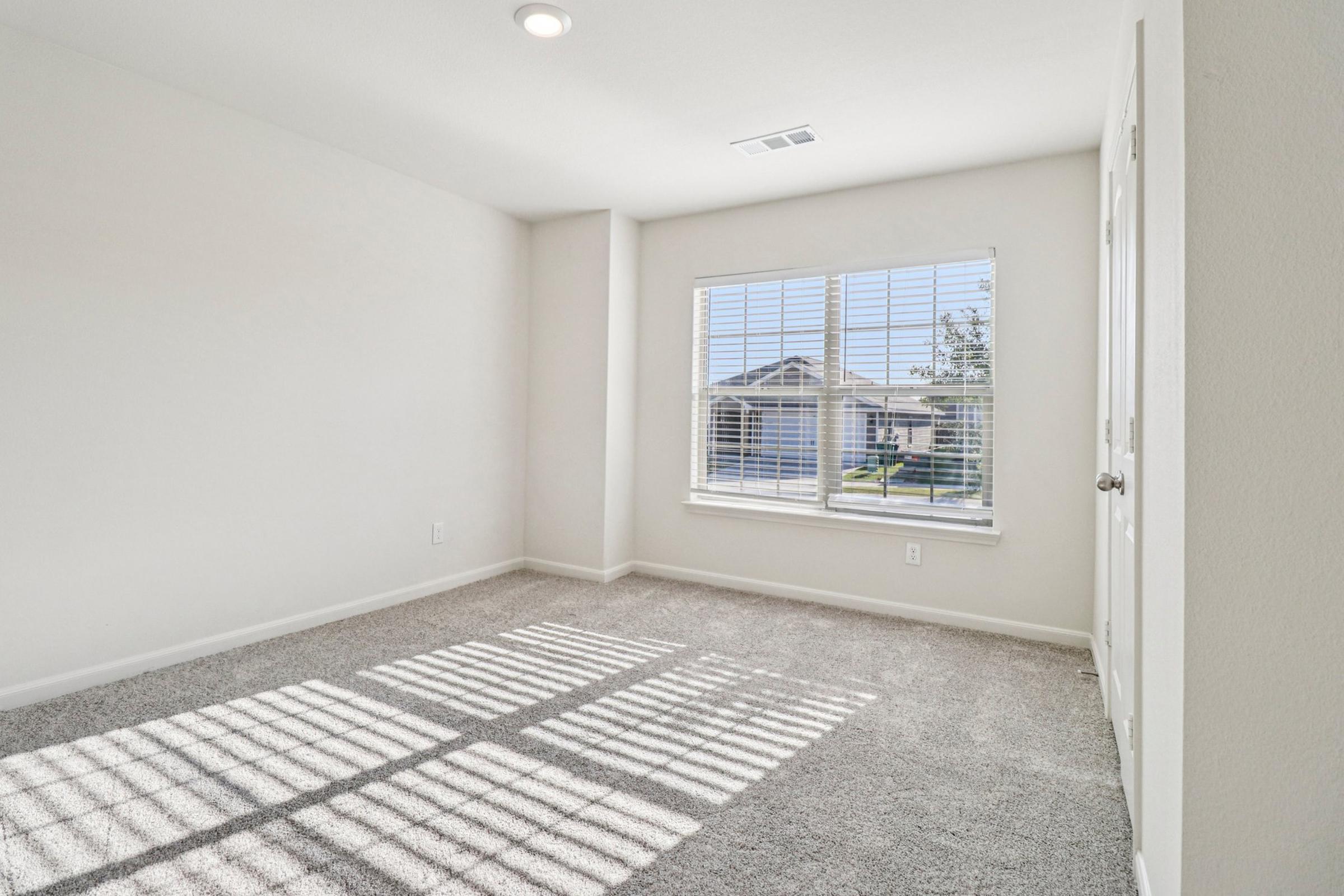 A well-lit empty room with beige walls and grey carpet. It features a large window with white blinds casting shadows on the floor. There is a door on the right side, and the overall appearance is clean and inviting, suitable for various uses.