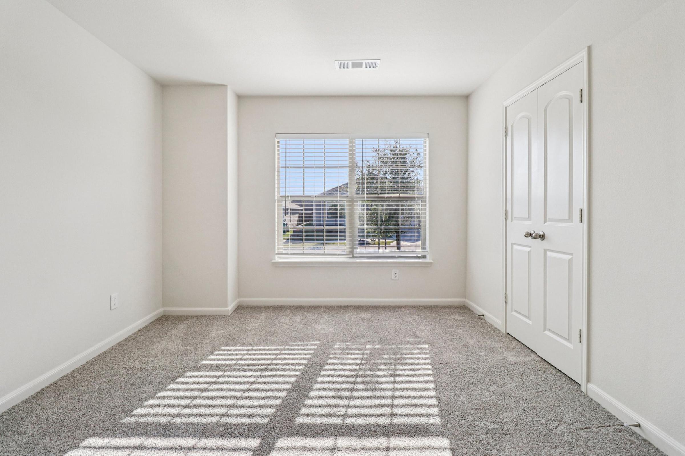 Empty room with light gray carpet and white walls. A large window with horizontal blinds allows natural light to enter, casting shadows on the floor. There is a door on the right side, and the overall space appears bright and airy, suggesting a freshly painted or newly constructed area.