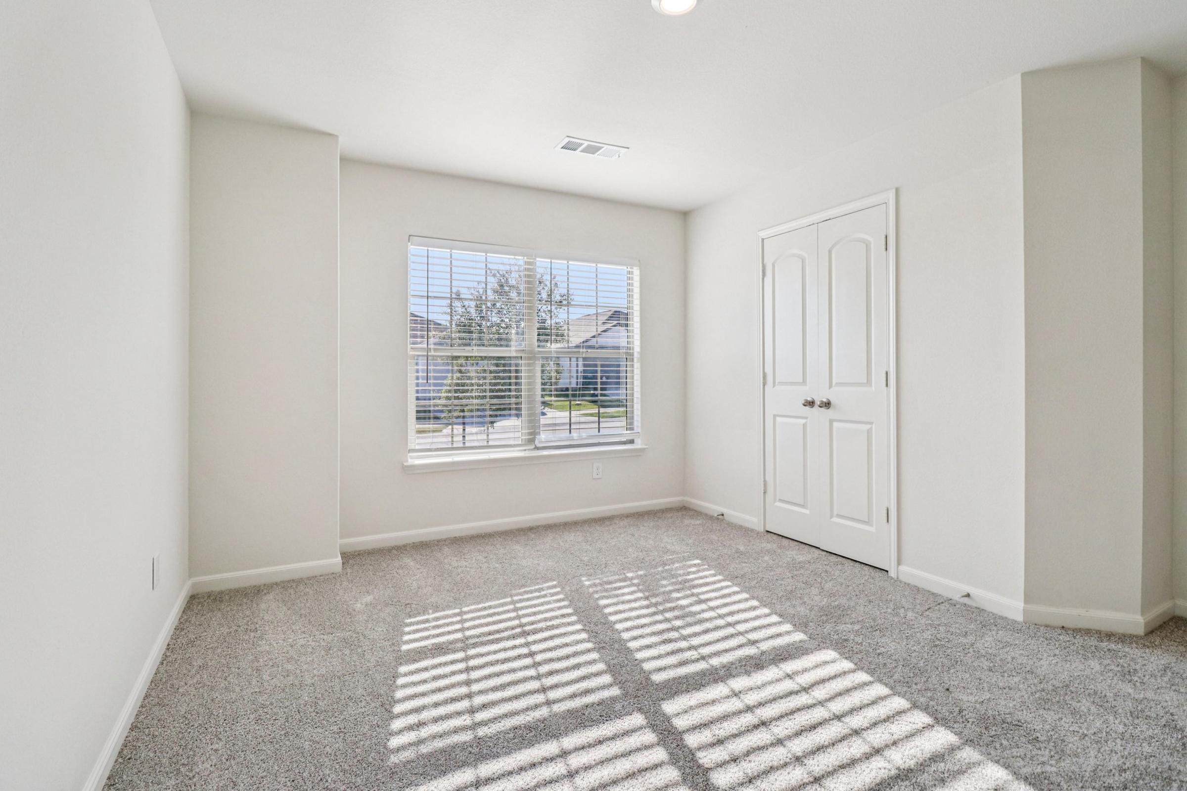 A bright, empty room with light gray carpet and white walls. There is a large window with blinds allowing natural light to fill the space, casting shadows on the floor. A double door closet is visible on one wall. The overall atmosphere is clean and inviting, ideal for personalization or decoration.