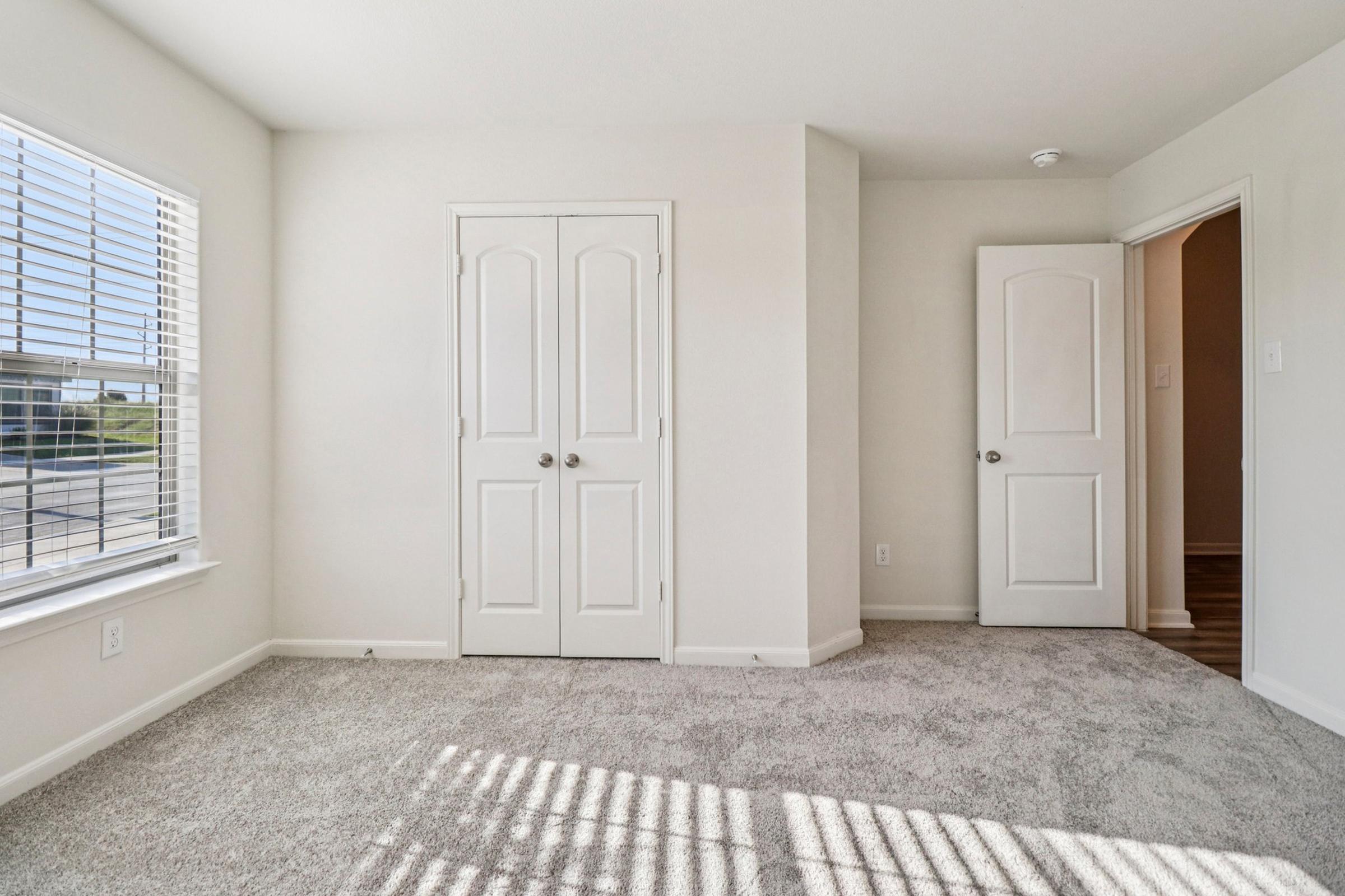 A bright, empty room with light-colored walls and carpet. Two windows with blinds allow natural light to enter. There are two doors: one leading to a closet and another to an adjoining room. The space appears clean and inviting, ideal for customization or furnishing.